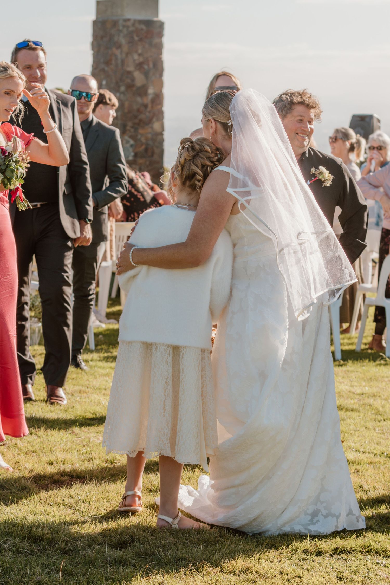 A bride in a flowing white dress and veil hugs someone in cream-colored attire during an outdoor wedding ceremony.