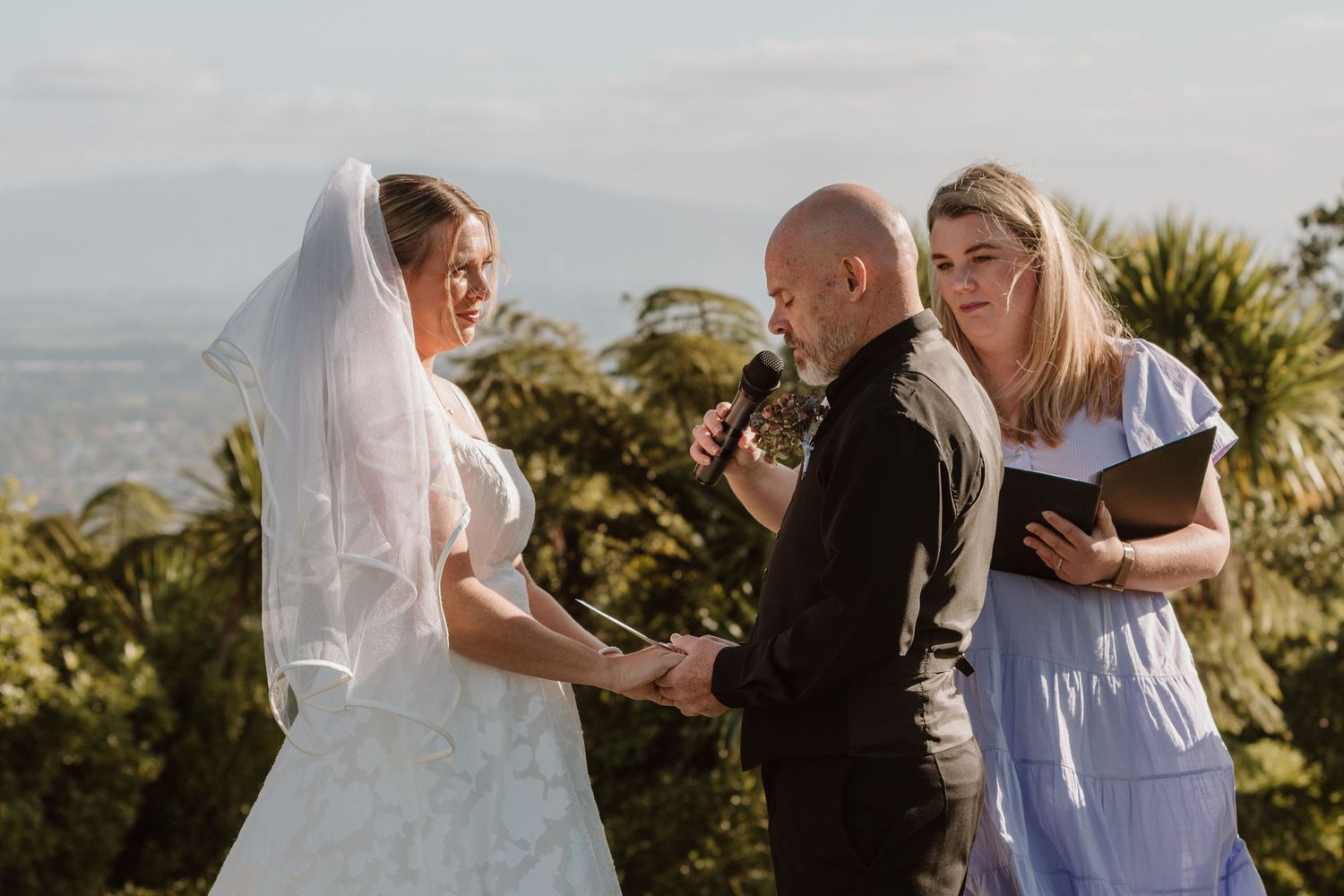 A seaside outdoor wedding ceremony shows a couple exchanging vows with an officiant on a sunny day.