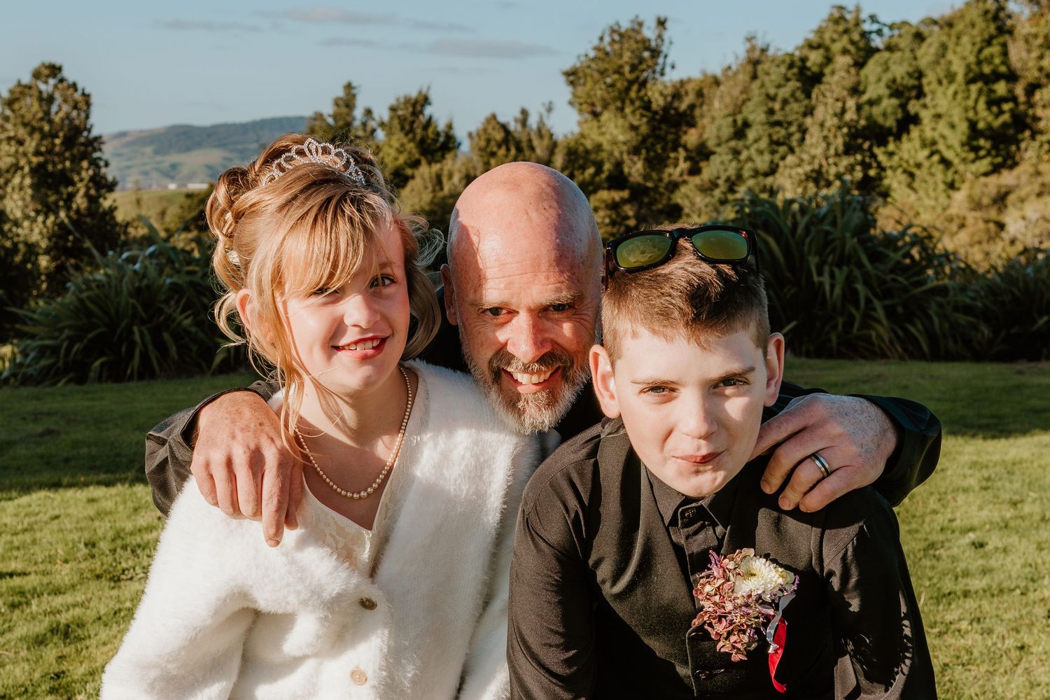 Three people share a warm embrace while smiling outdoors on a sunny day with green hills and trees in the background.