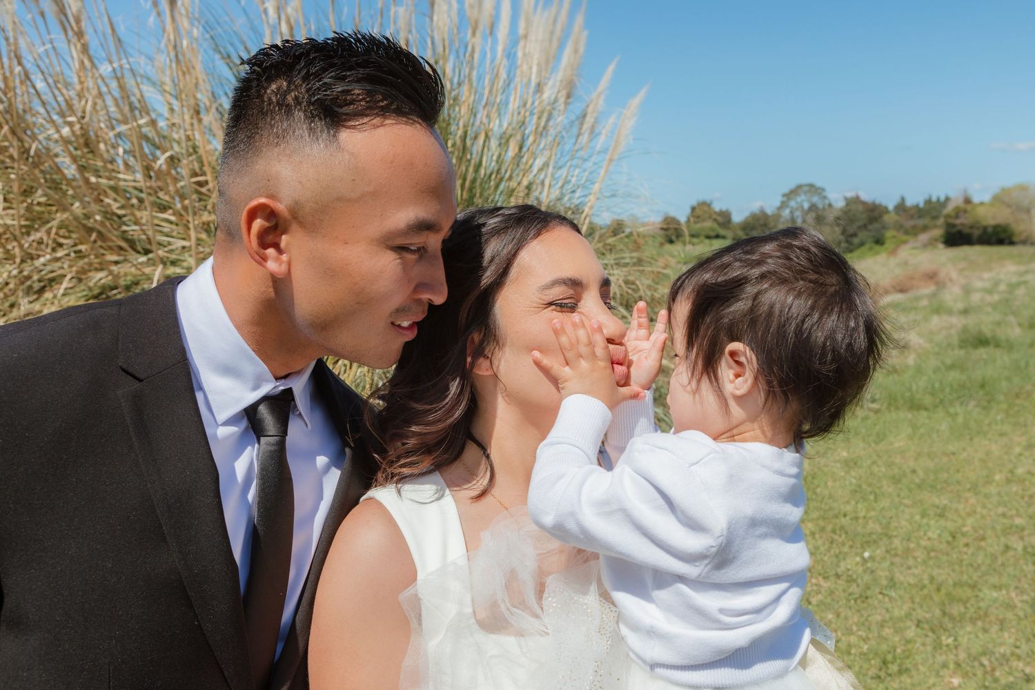 A happy family shares a tender moment together on a sunny day in a grassy field with tall reeds in the background.