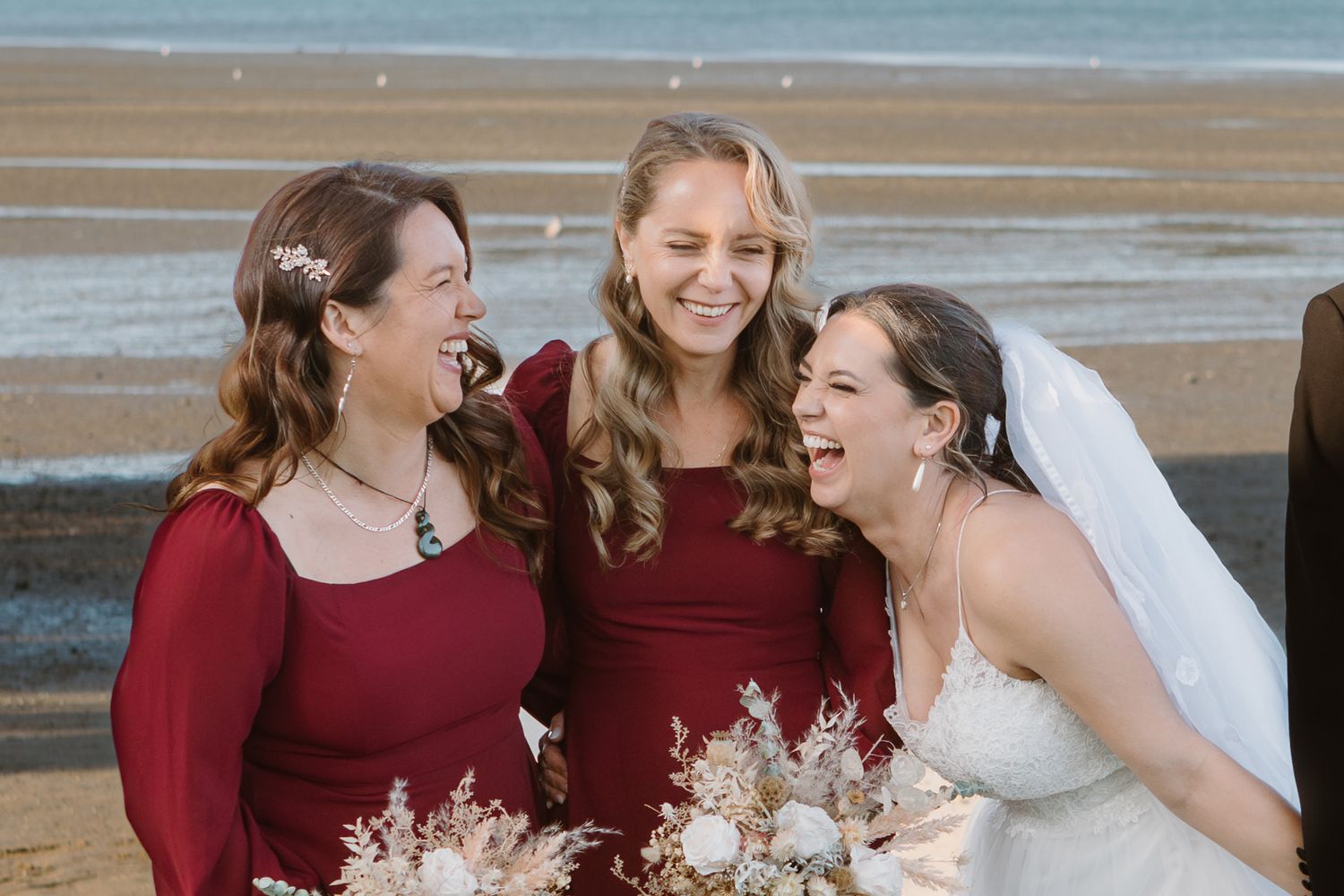 Five joyful people in burgundy bridesmaid dresses and one in white share candid moments on a sunny beach.