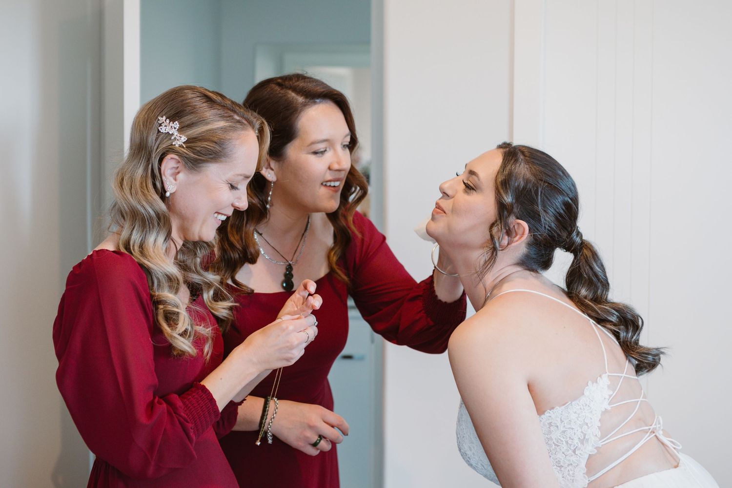 A bride shares joyful moments with her bridesmaids wearing burgundy dresses during wedding day preparations.