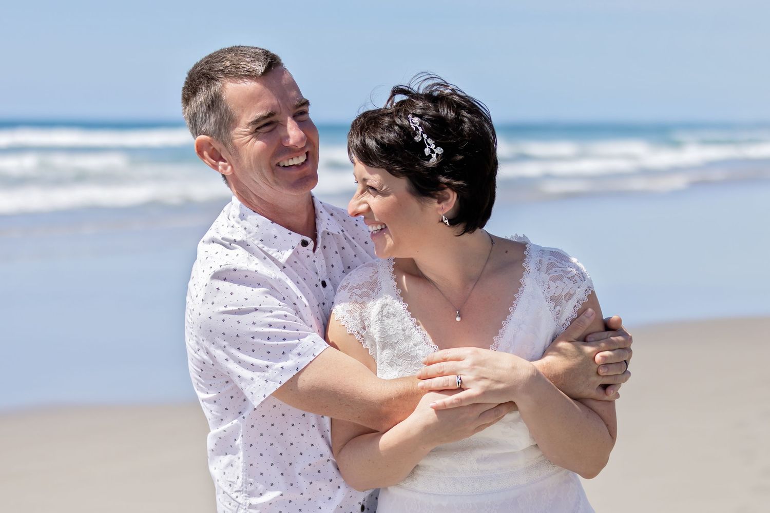 A happy couple embrace and laugh together on a sunny beach with ocean waves in the background.