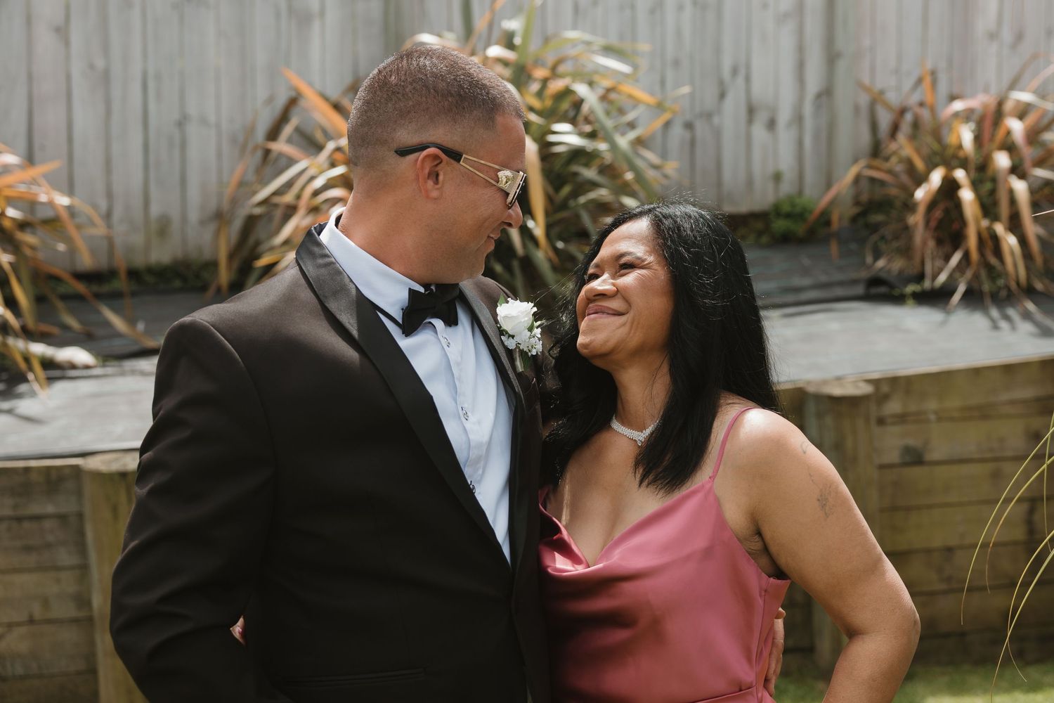 A couple shares an intimate moment in formal attire at an outdoor garden setting with a wooden fence backdrop.