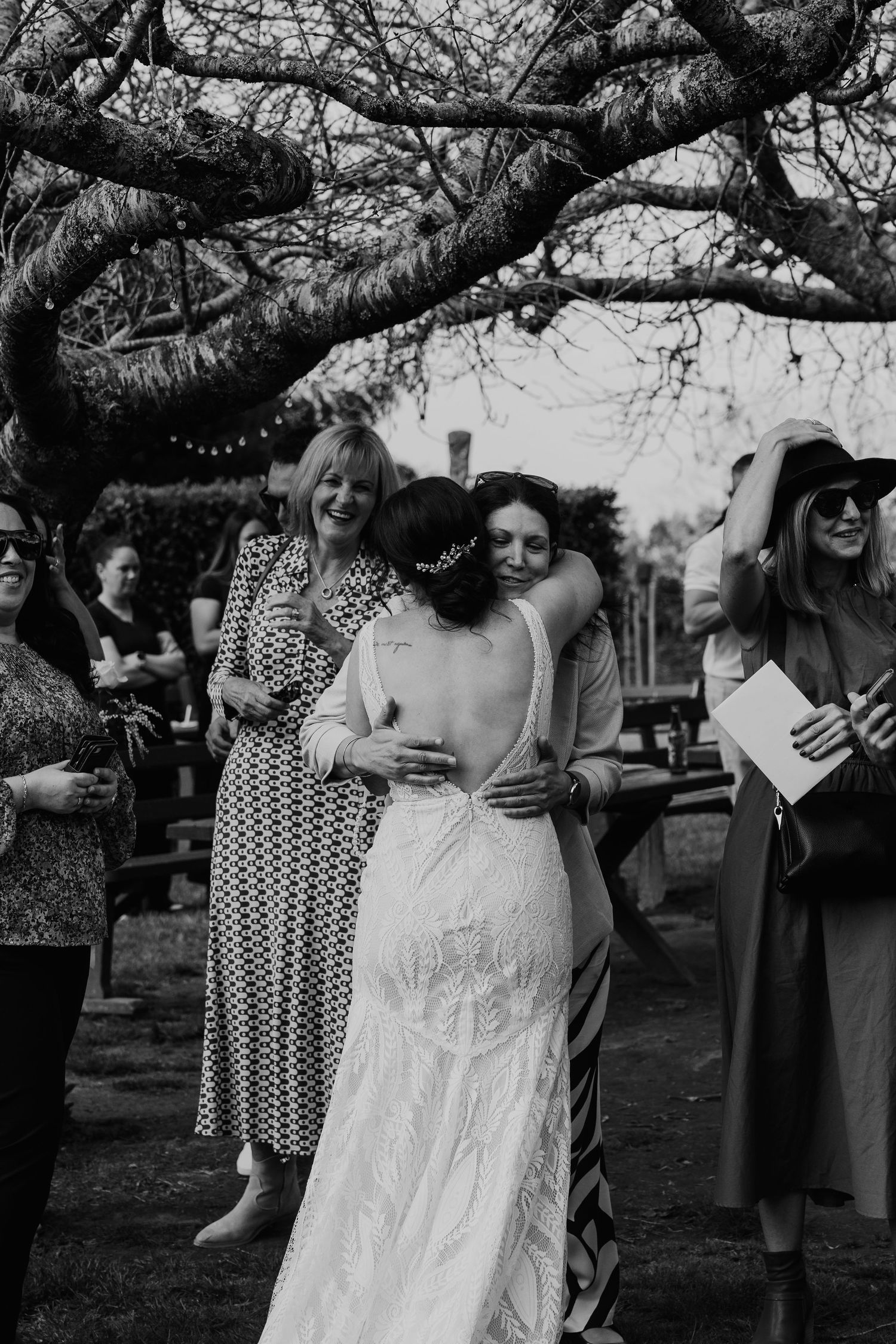 Black and white photo of a casual outdoor wedding gathering under a tree with guests in vintage-style dresses.
