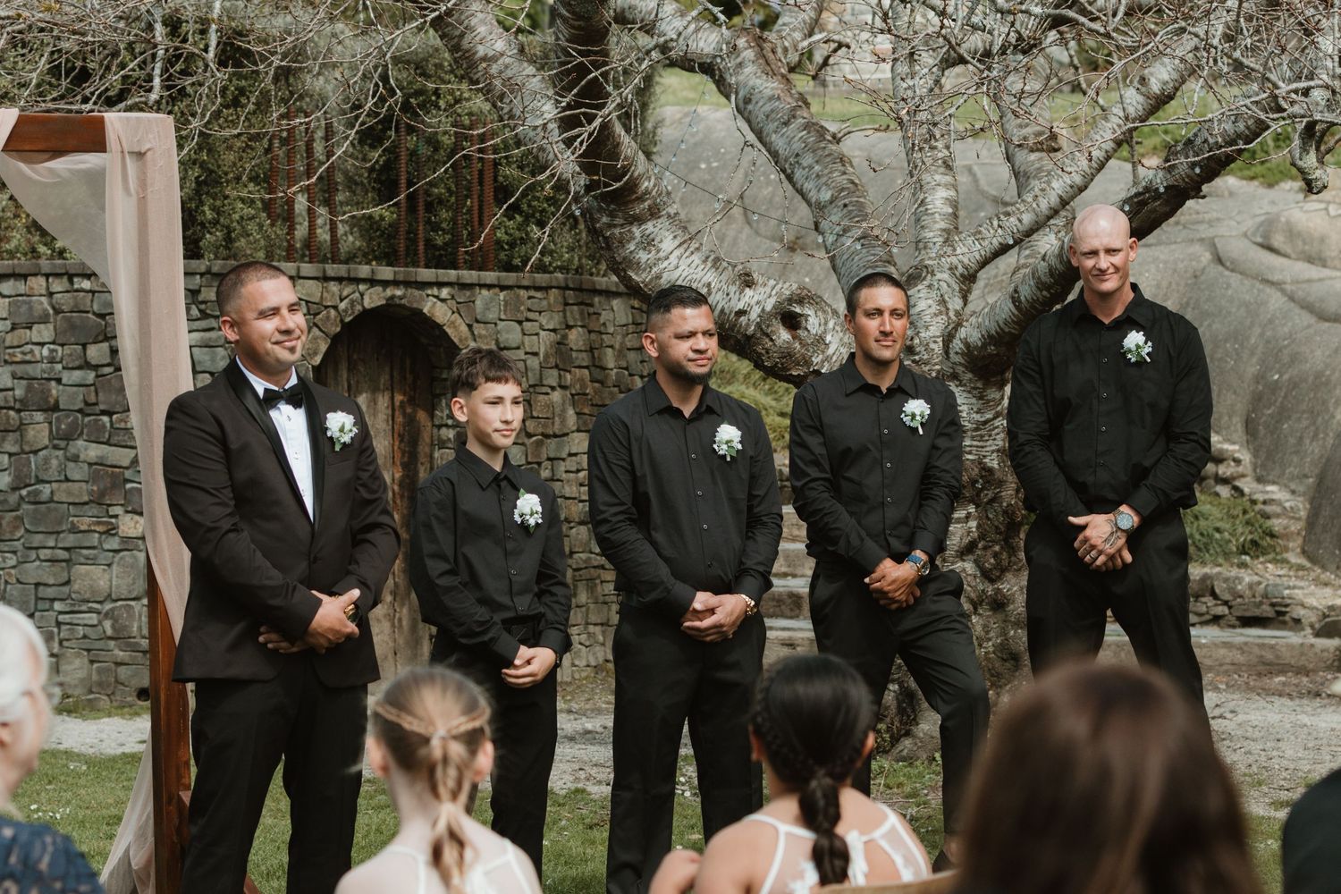 A group of groomsmen in black suits share a candid moment of laughter during a wedding celebration outdoors.