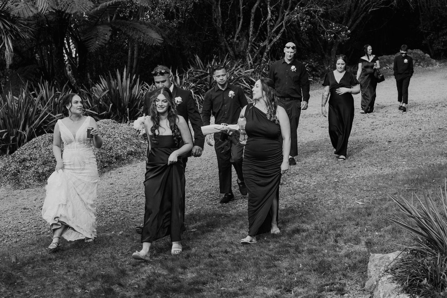 Wedding party walks in a line along a garden path during an outdoor black and white ceremony.
