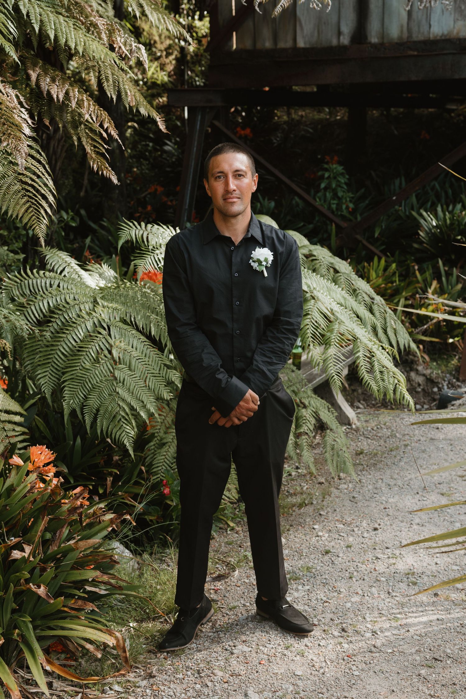 A figure in dark clothing stands on a gravel path surrounded by lush ferns and tropical plants in a garden setting.