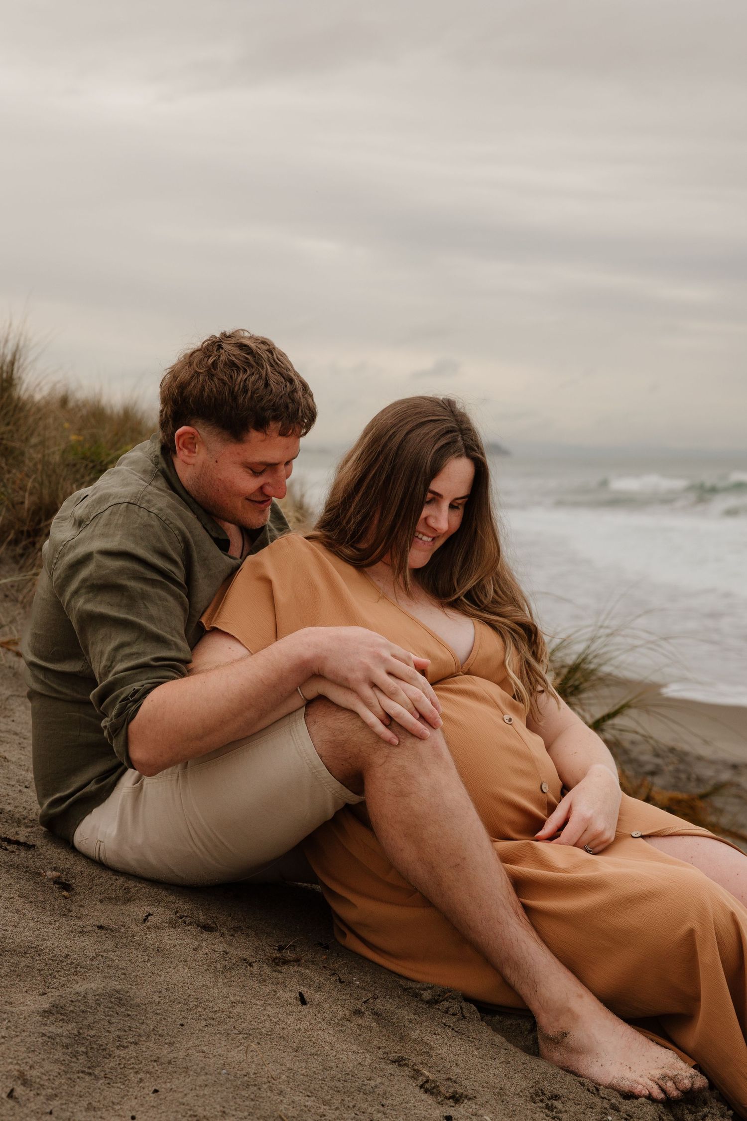 A couple in casual attire embraces intimately on a sandy beach with cloudy skies and ocean waves in the background.