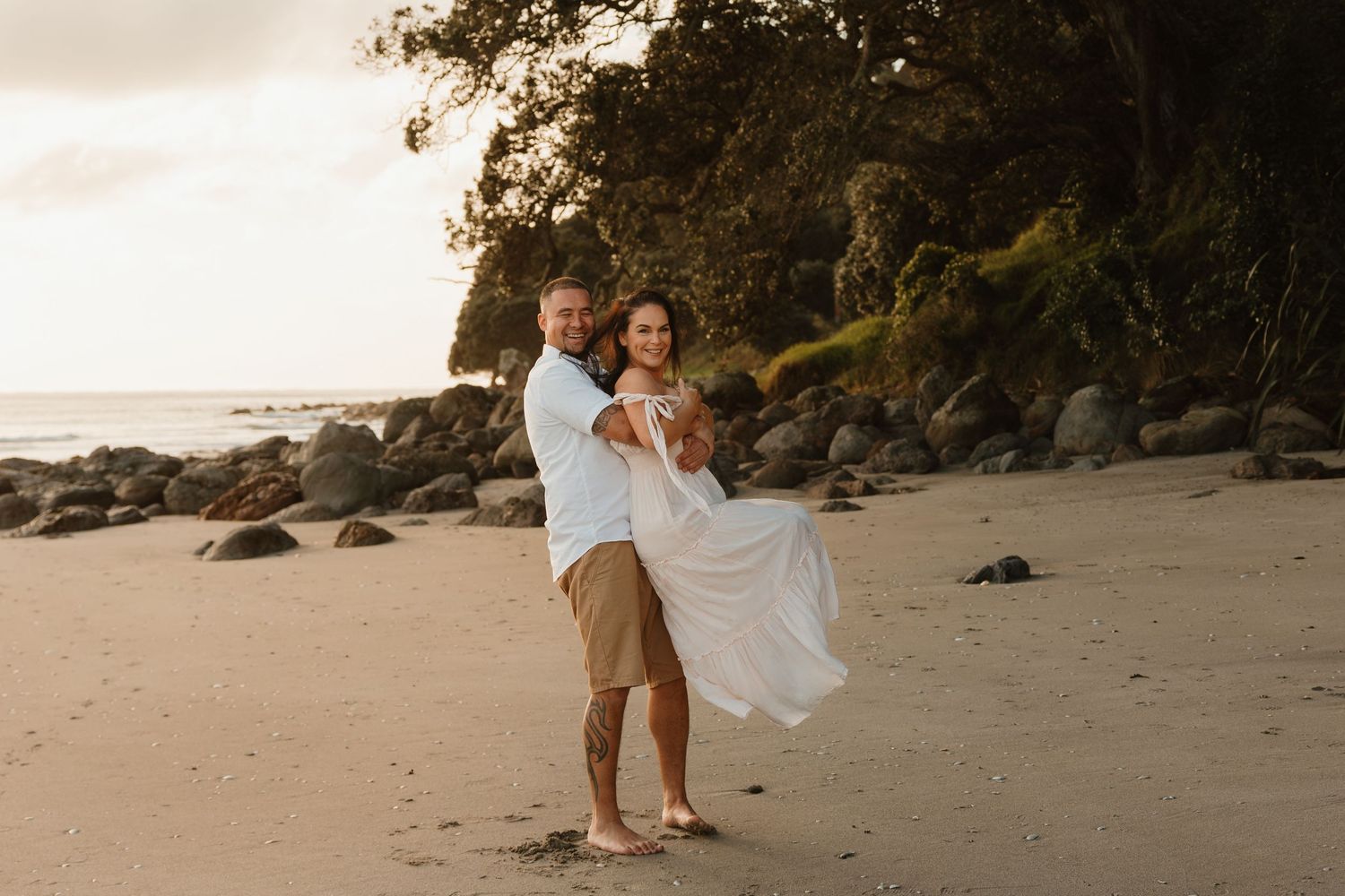 Couple walks hand in hand along scenic beach with rocky cliffs and golden sand at sunset.