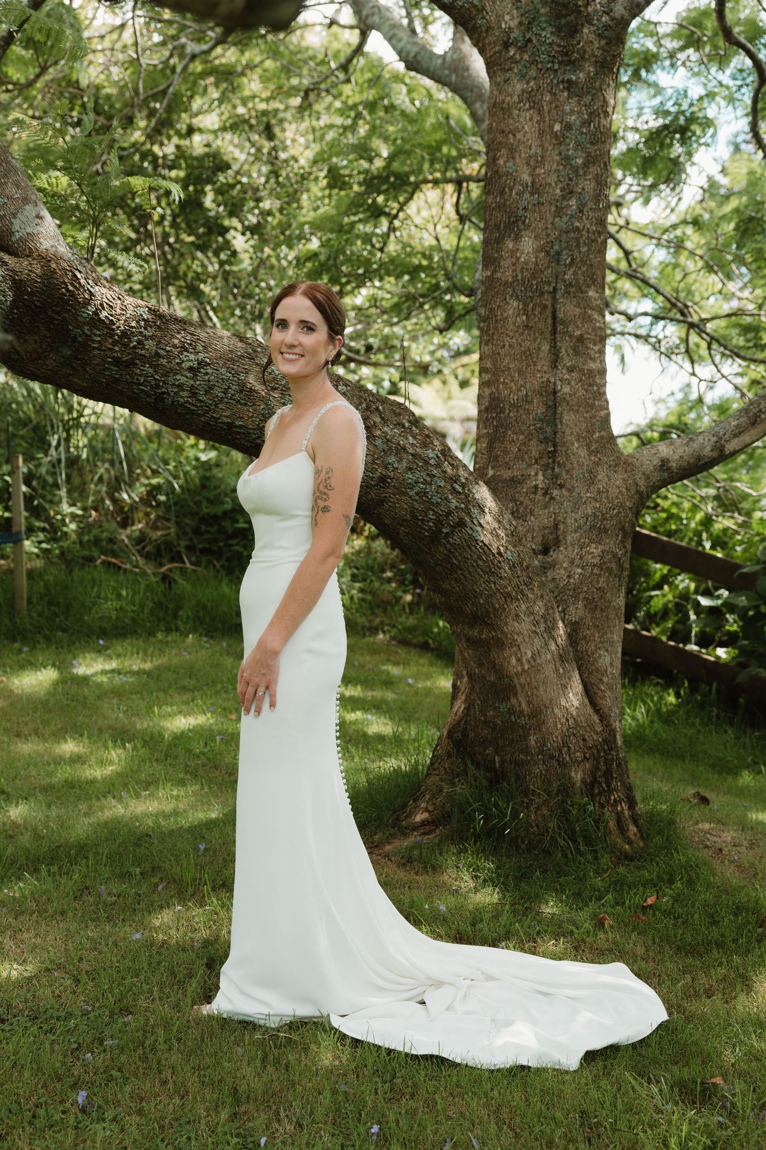 Bride in white satin gown with train poses gracefully beside a large tree in a lush green garden.