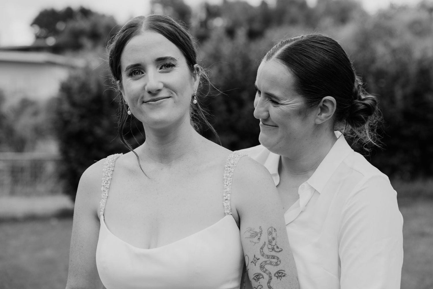 Black and white photo of two women smiling closely together outdoors, one in a white dress with a floral tattoo.