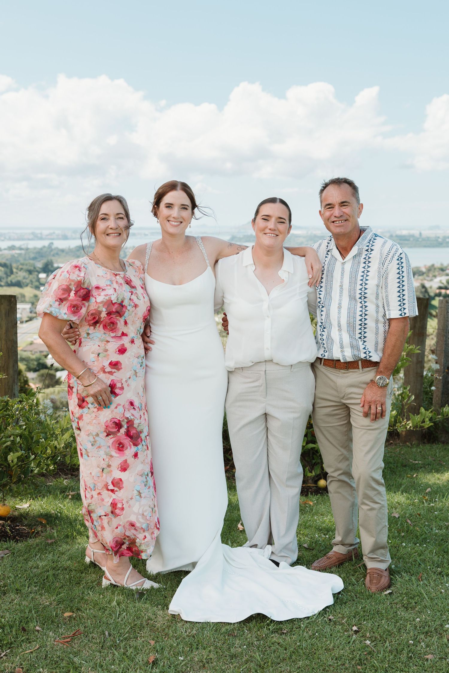 Bride in white gown poses with family of three outdoors at scenic wedding venue under blue sky.