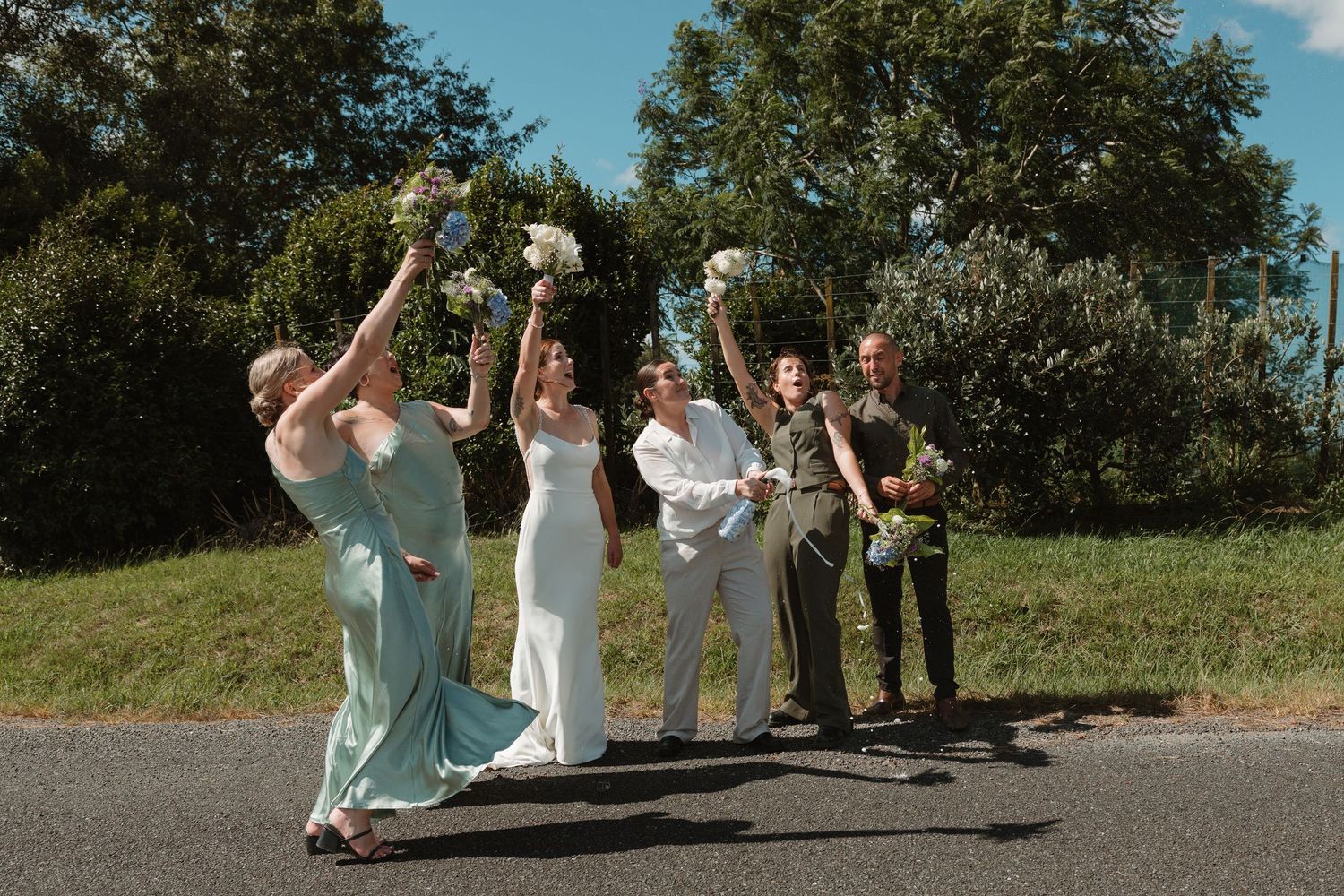 Wedding party celebrating outdoors, raising bouquets joyfully on a road, bridesmaids in sage green and couple in white and gray.