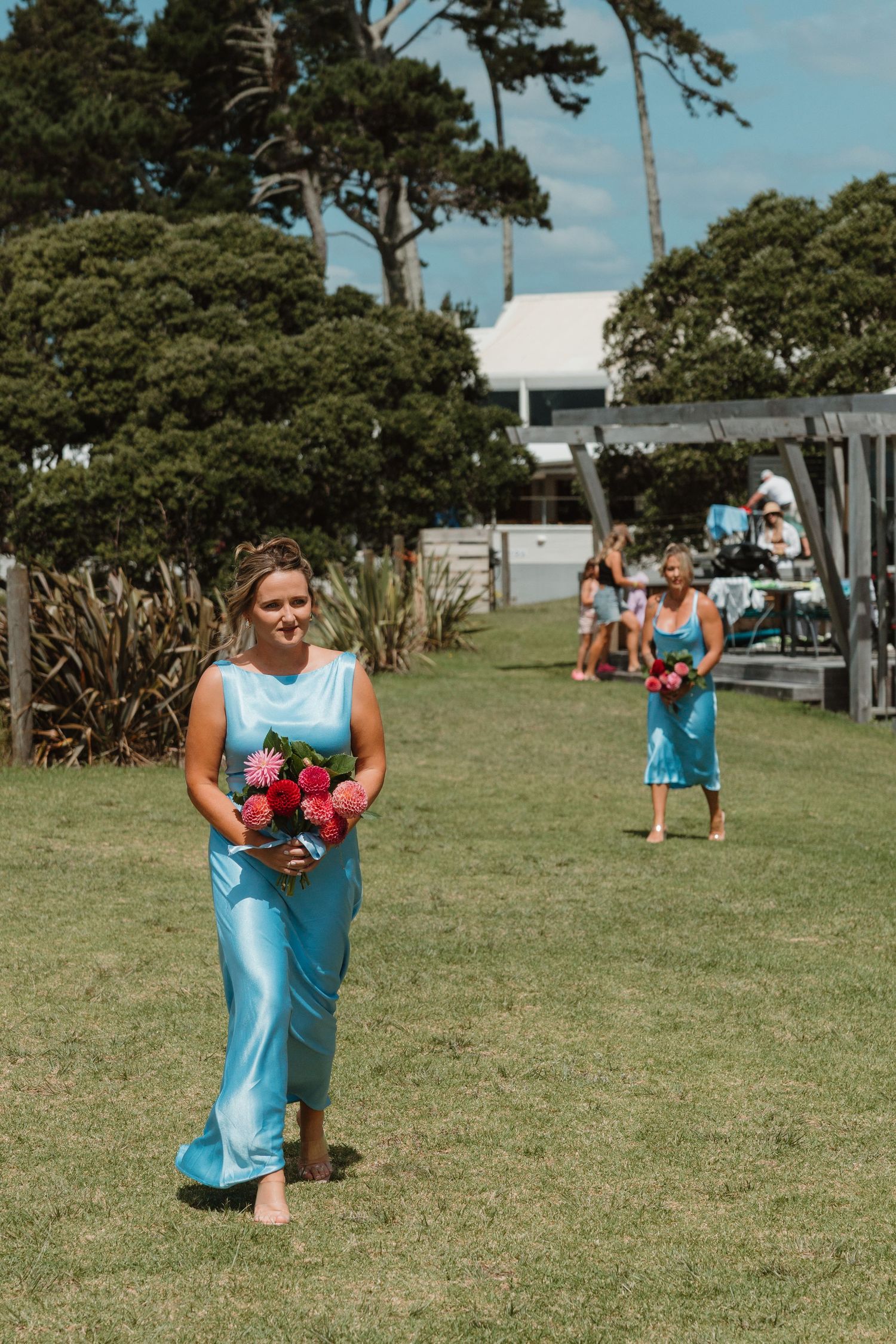 Bridesmaids in turquoise satin dresses carrying pink bouquets walking across green lawn at outdoor wedding ceremony.