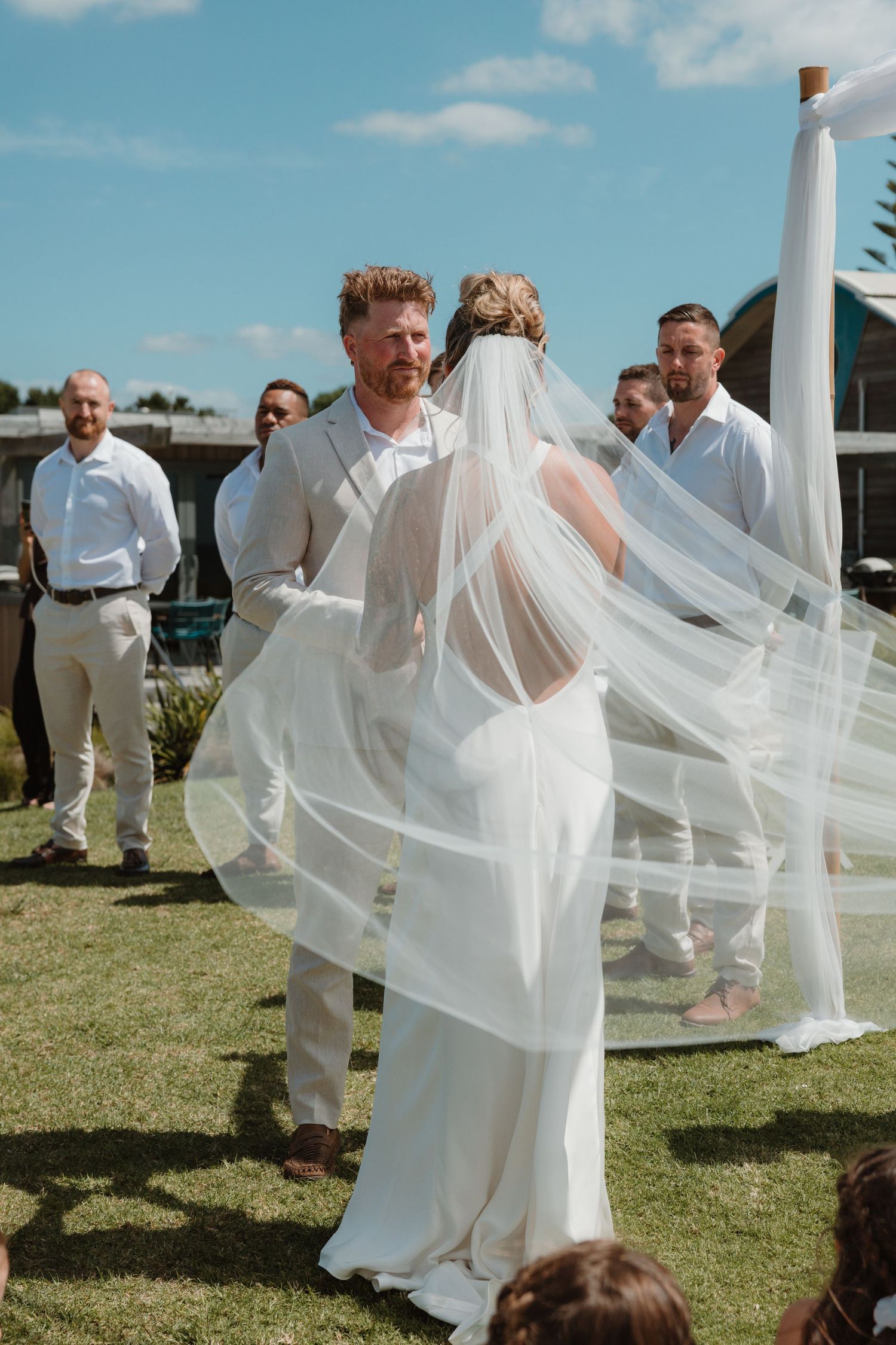 Bride and groom exchange vows outdoors, her veil flowing in the breeze, groomsmen in white shirts behind them.