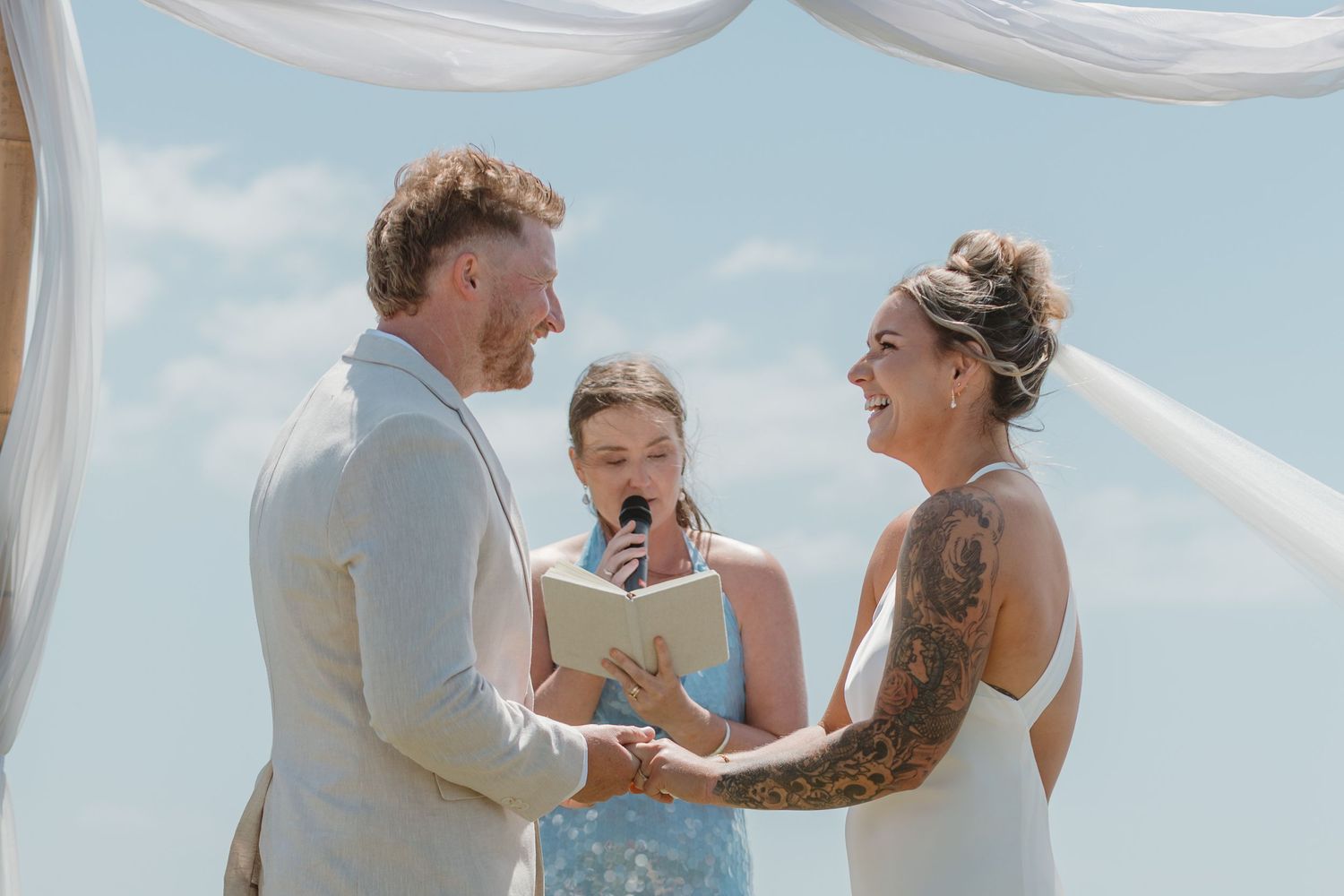Couple exchanging vows at outdoor beach wedding ceremony with officiant and flowing white fabric backdrop.