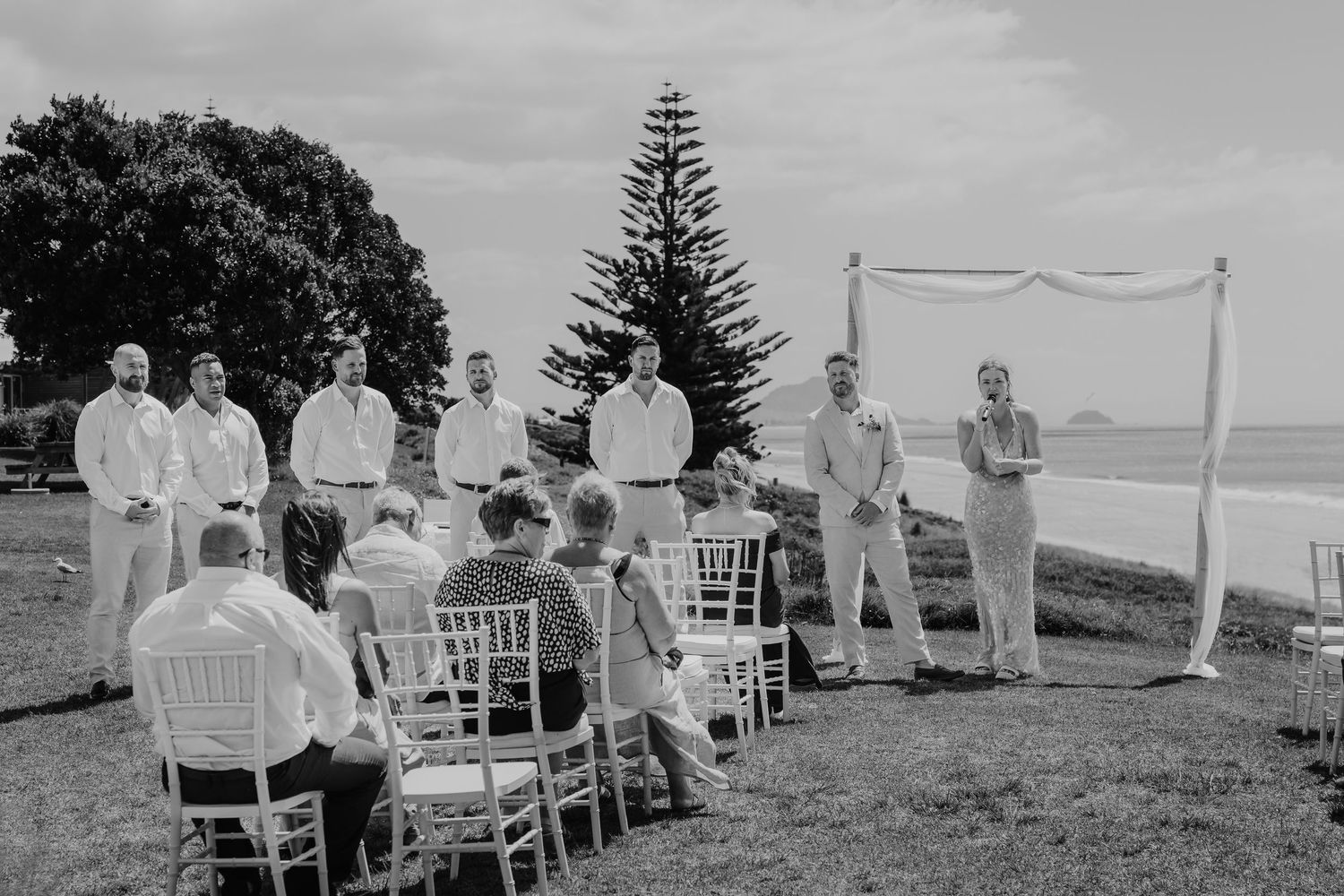 Black and white photo of a wedding ceremony outdoors with groomsmen standing near a decorated arch by the ocean.