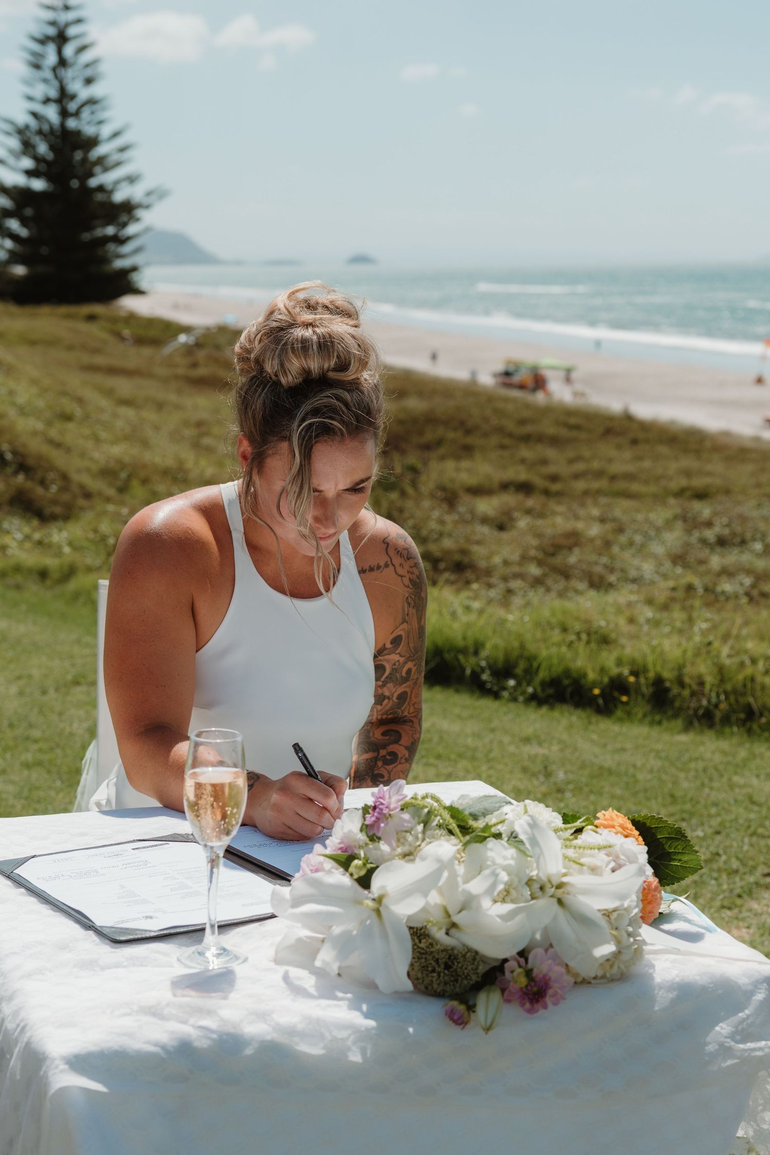 Bride signing wedding register at outdoor beachside table with champagne and white floral bouquet, tattoos visible.