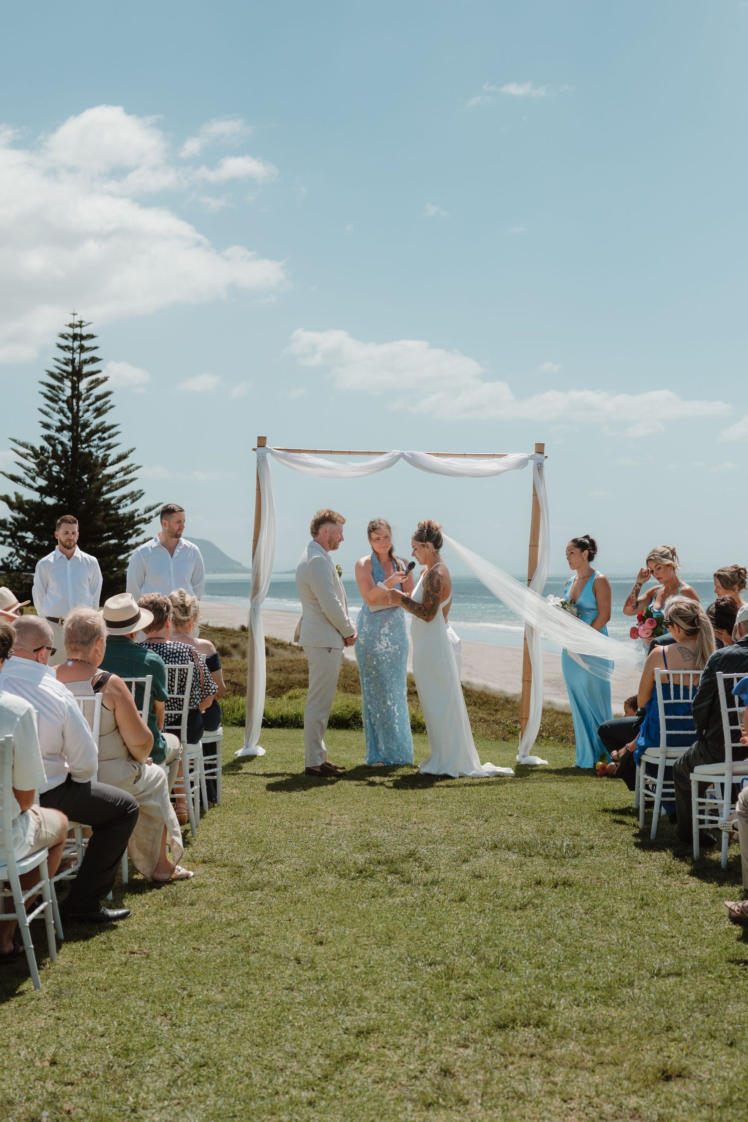 Outdoor beach wedding ceremony with couple at floral arch, guests seated on white chairs, ocean backdrop on sunny day.