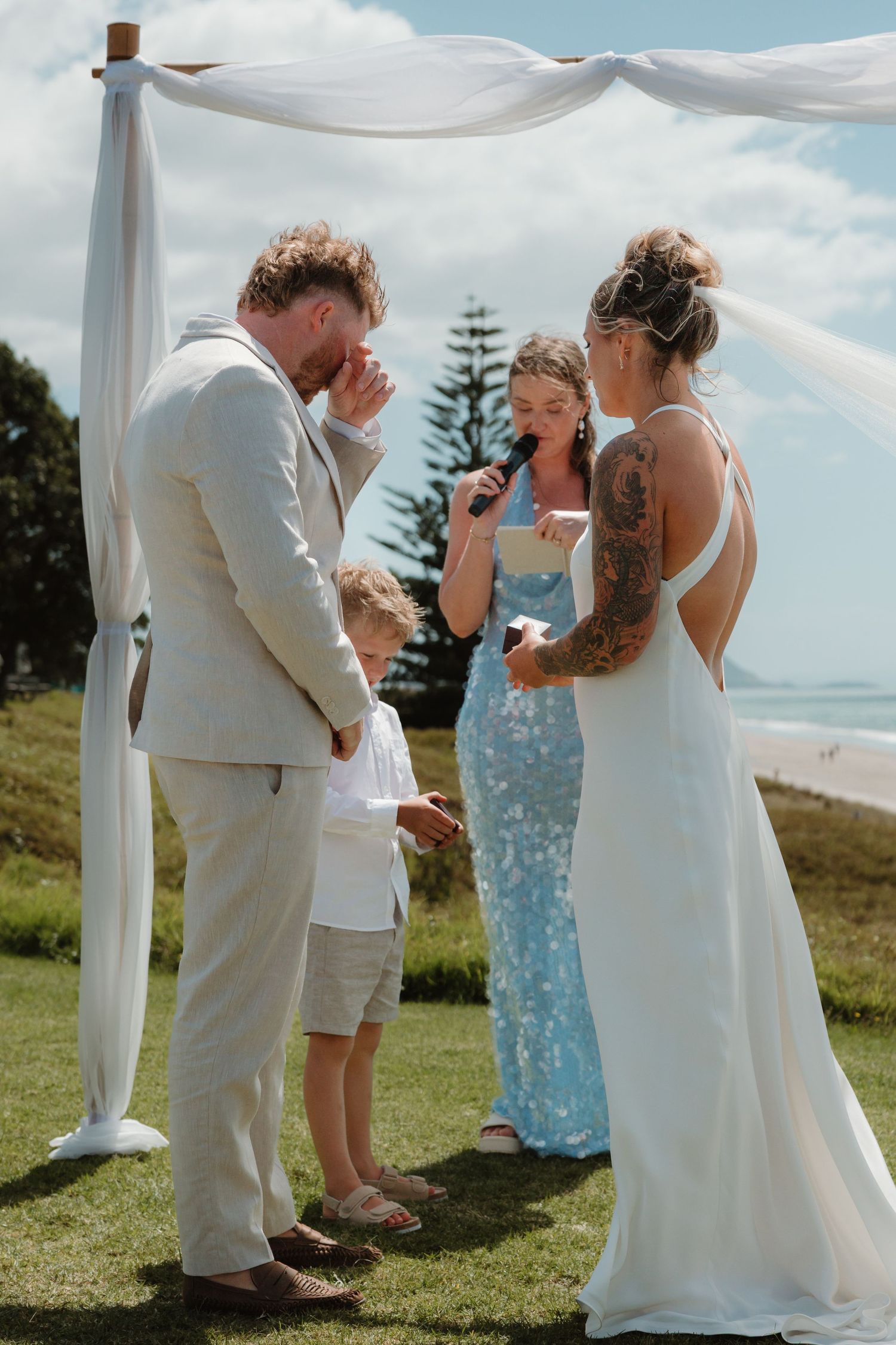 Emotional outdoor wedding ceremony by the beach, groom wiping tears as bride reads vows, officiant in blue dress.