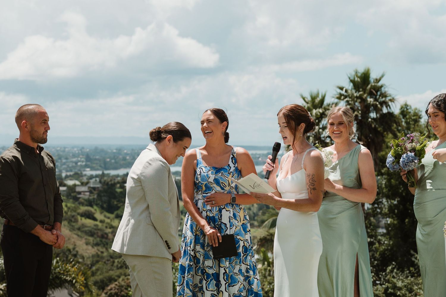 Outdoor wedding ceremony with bride and guests laughing on a hilltop overlooking a city skyline with palm trees.