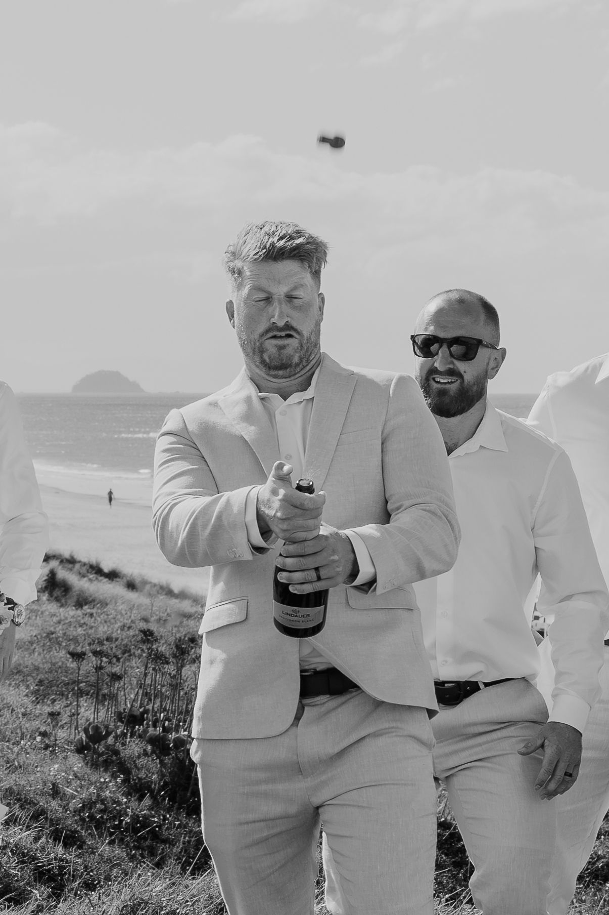 Groom opening champagne bottle with groomsmen in light suits at outdoor beach wedding celebration.