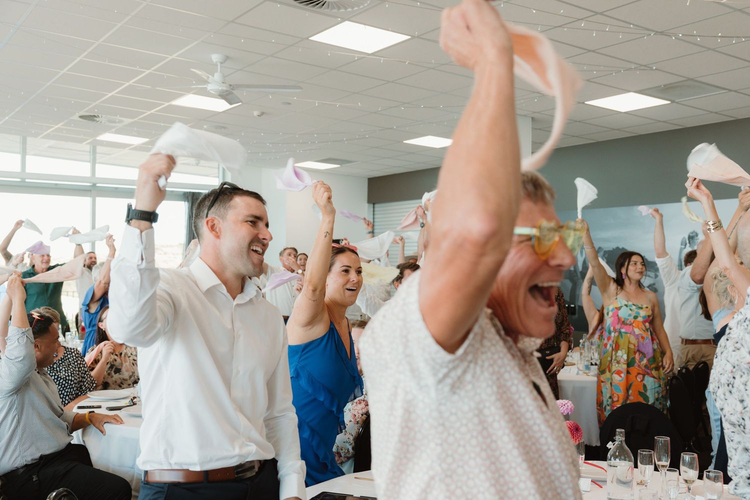 Excited guests wave napkins and cheer at a lively indoor celebration or wedding reception event.