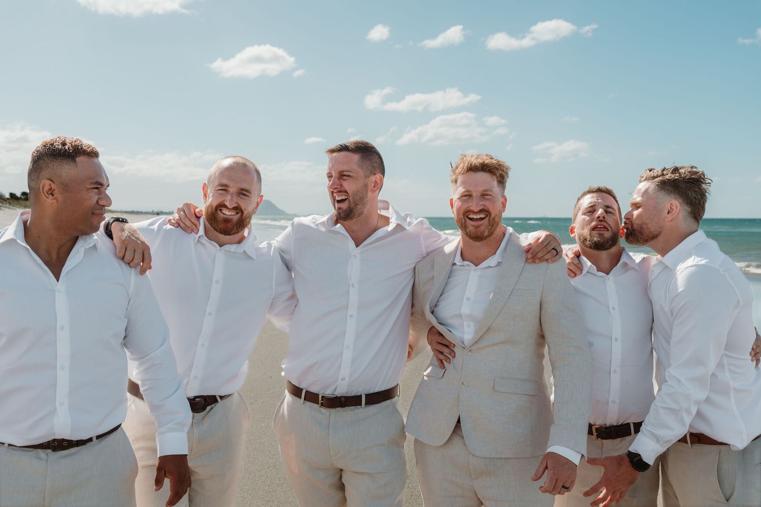 Six groomsmen in matching beige suits laugh together on a sunny beach with blue ocean waves in the background.