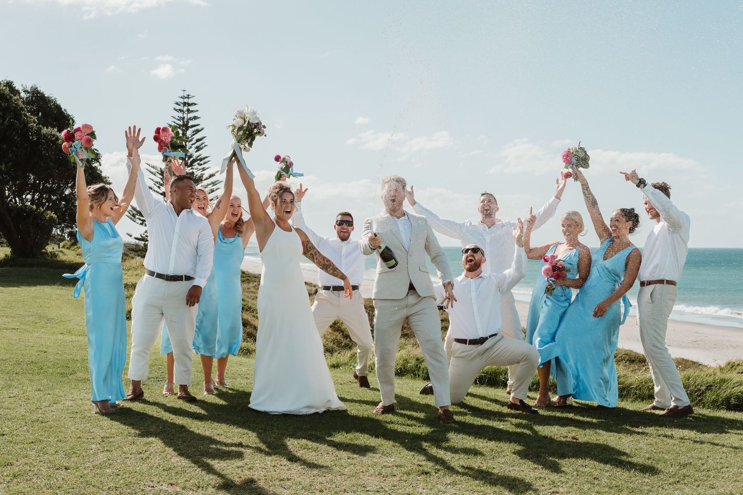 Wedding party celebrating outdoors near beach, bridesmaids in turquoise dresses, groomsmen in white, raising bouquets joyfully.
