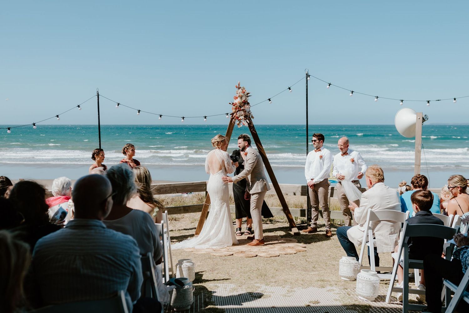 Panoramic view of a beach wedding setup with string lights and ocean backdrop. Papamoa Surf Lifesaving Club Wedding