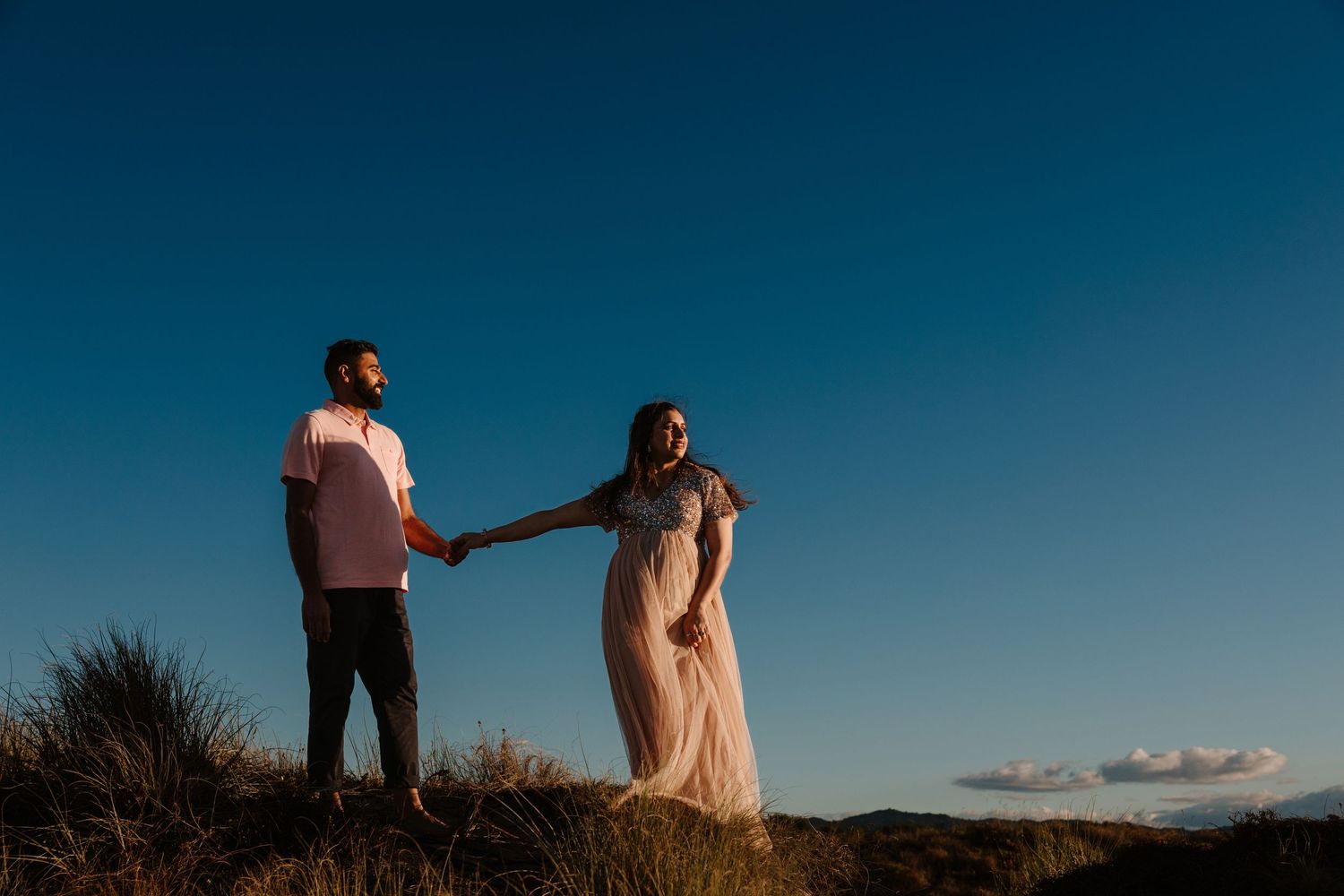 Two silhouetted figures hold hands on a hilltop against a deep blue sky at sunset, during a beach maternity photoshoot in Tauranga