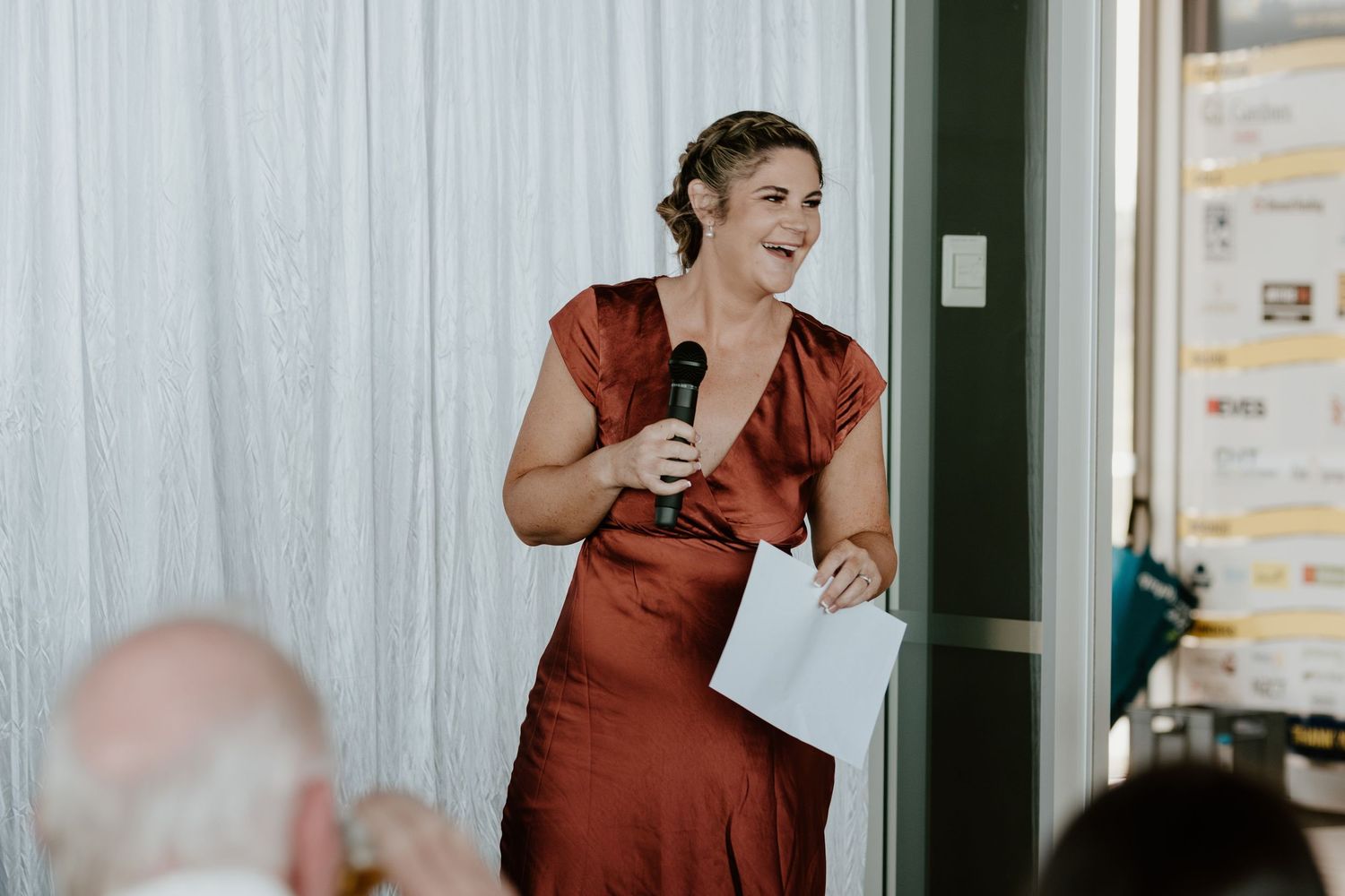 A person gives a cheerful wedding speech while wearing a rust-colored dress and holding a microphone.