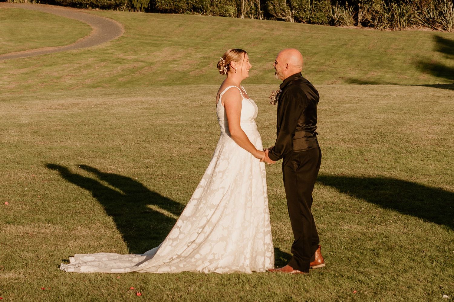 Two people in formal wedding attire sharing a tender moment on a sunlit grassy field.