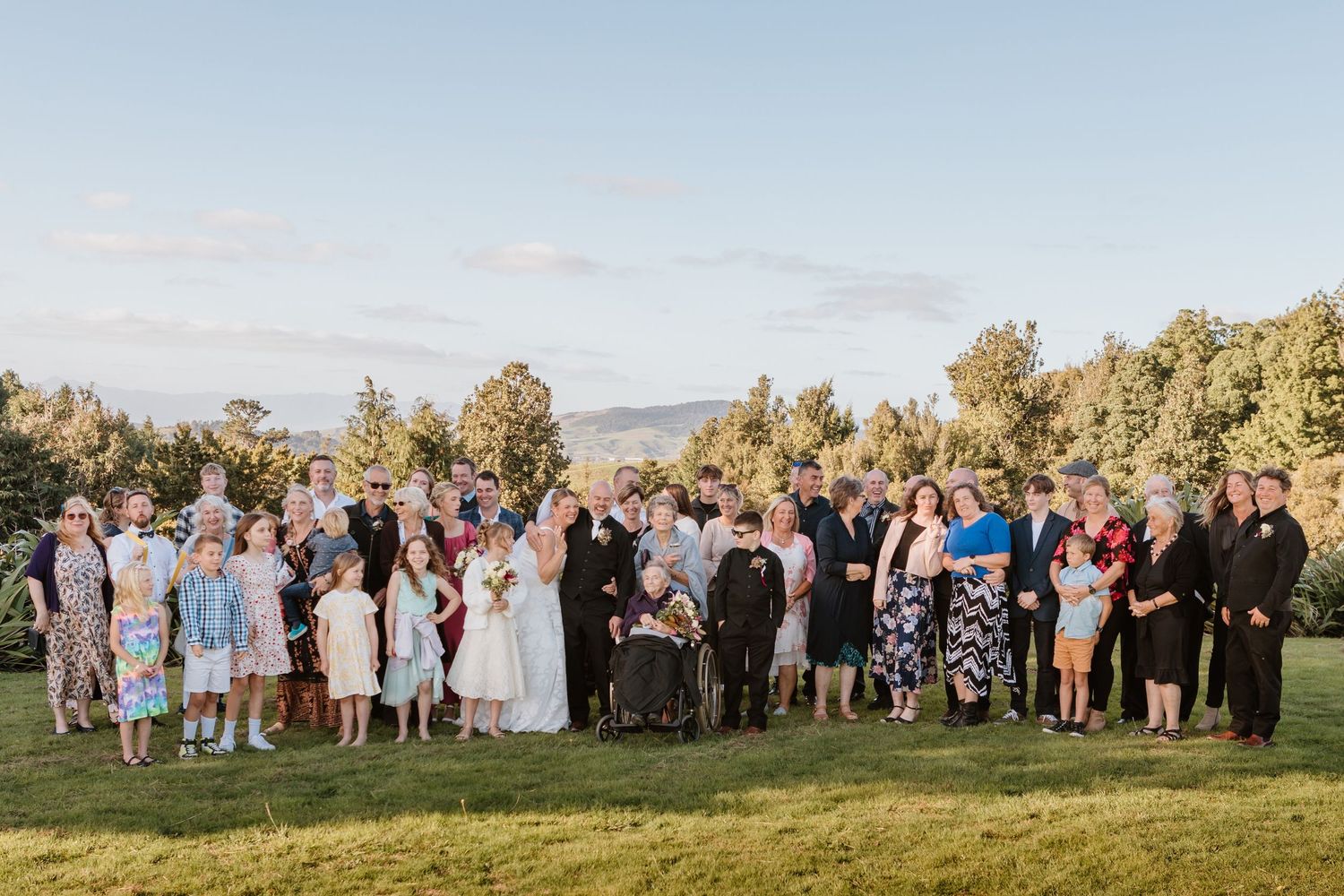 Wedding celebration group portrait captured outdoors on hilltop with mountain landscape at dusk.