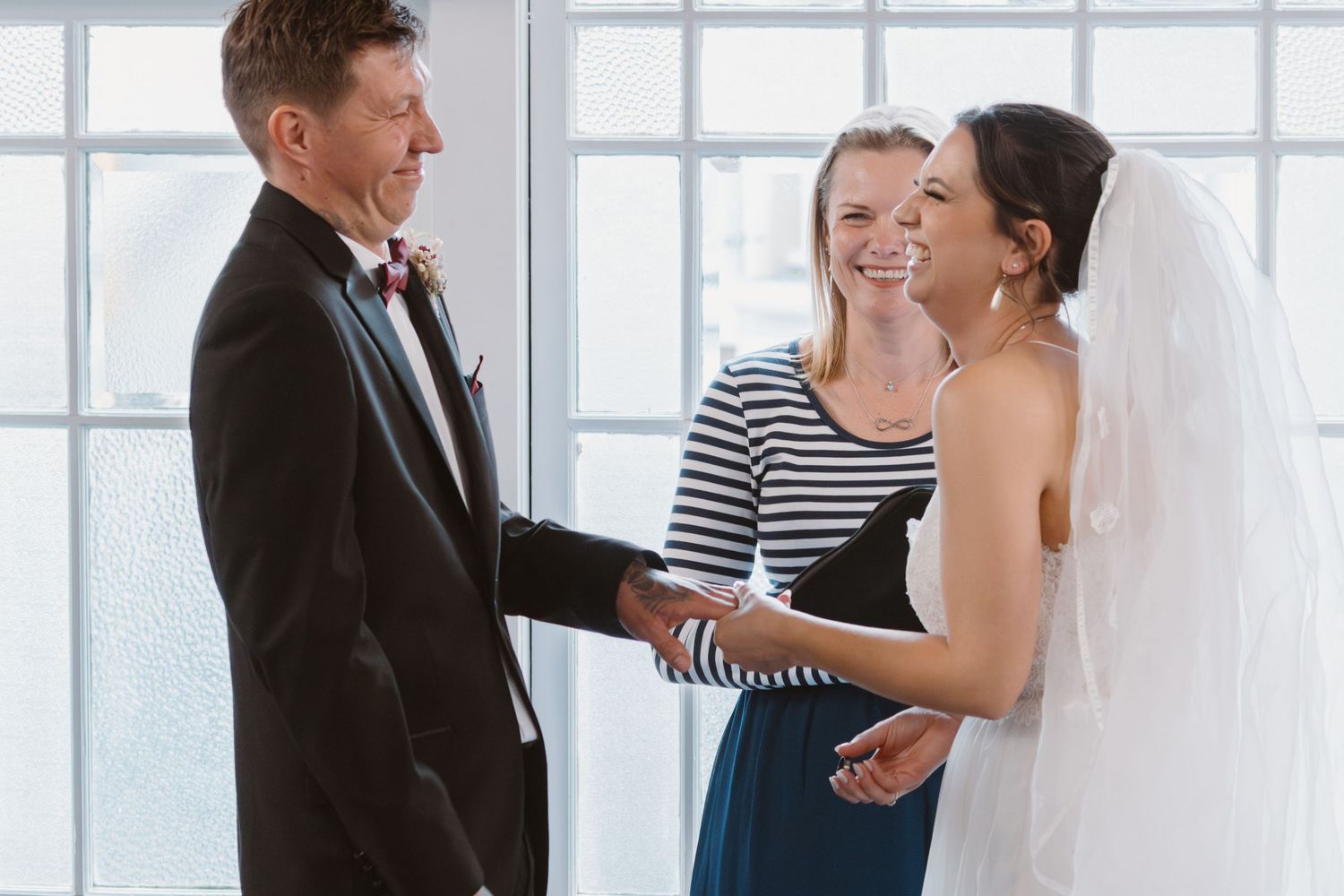 Wedding party laughs together in a bright, sunlit room with large windows in the background.