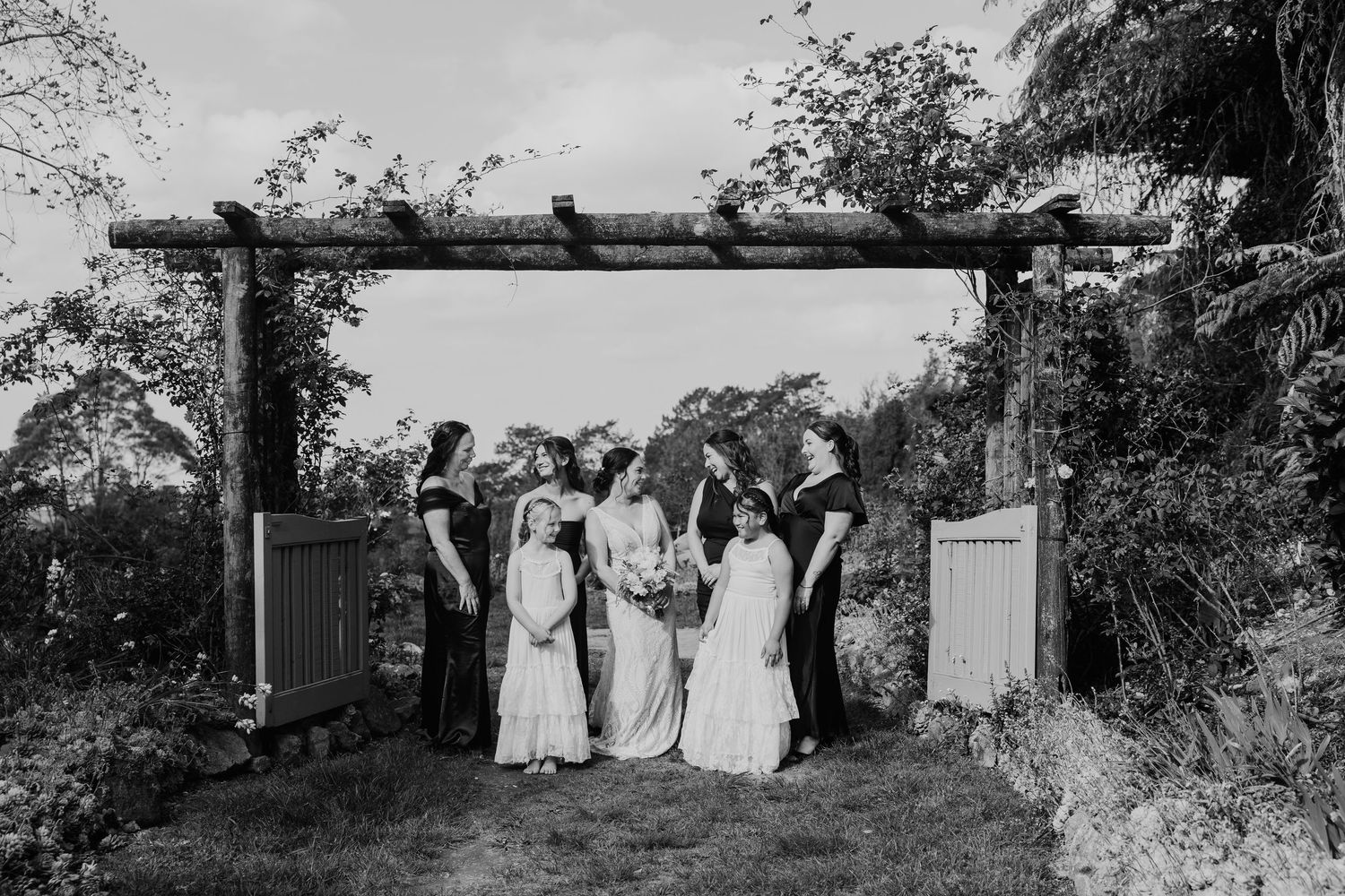 A black and white photograph of a wedding party standing under a rustic wooden archway in a garden setting.