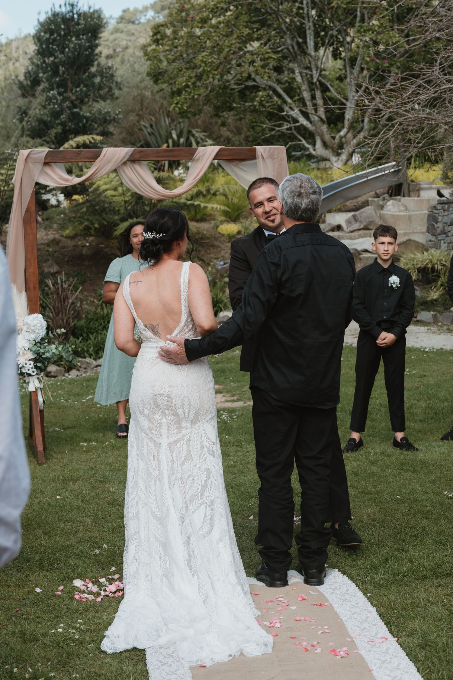A couple shares a playful moment during their outdoor wedding ceremony with pink petals scattered on the grass.