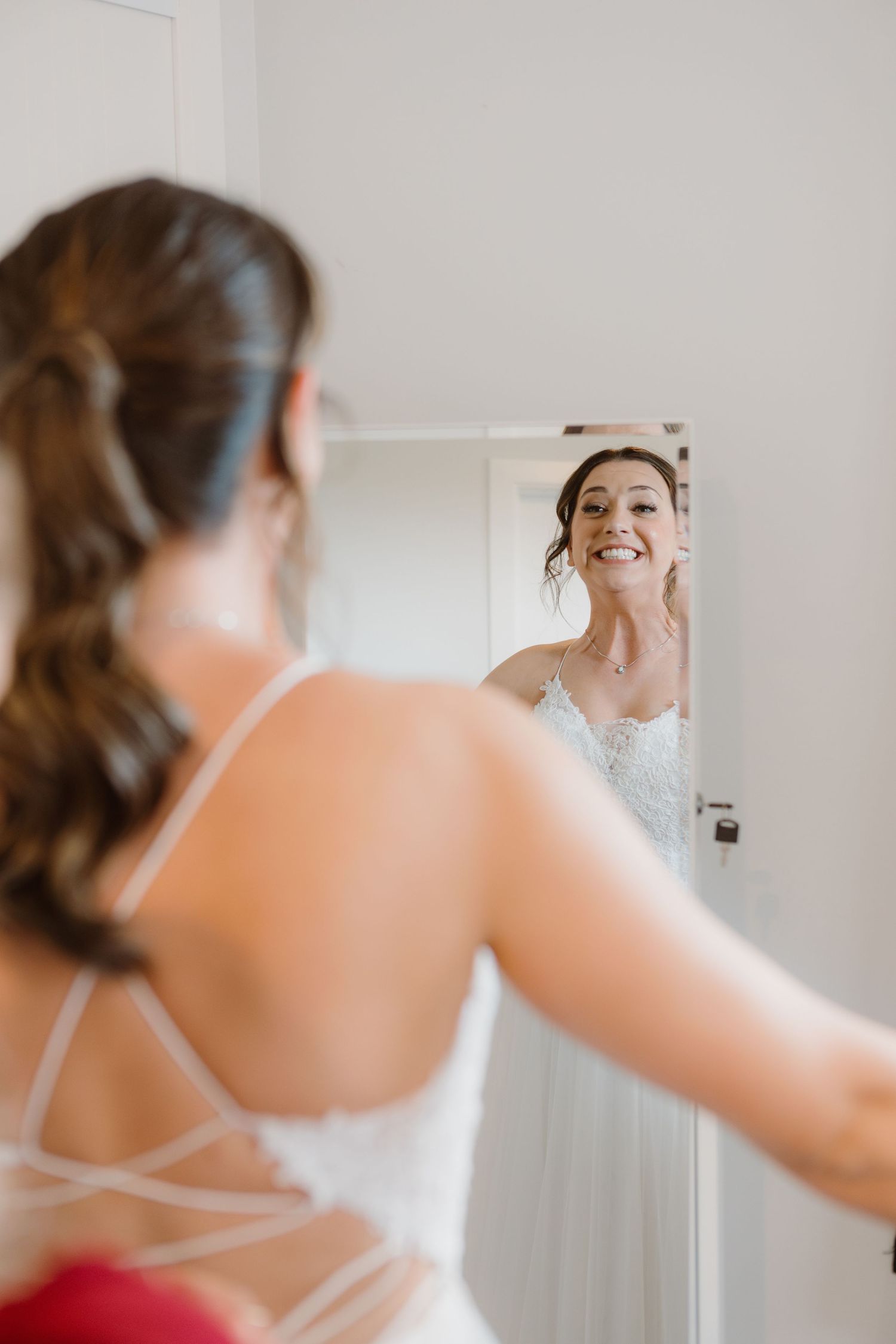 Bride in white dress looking at her reflection in mirror with joyful expression during wedding preparations.