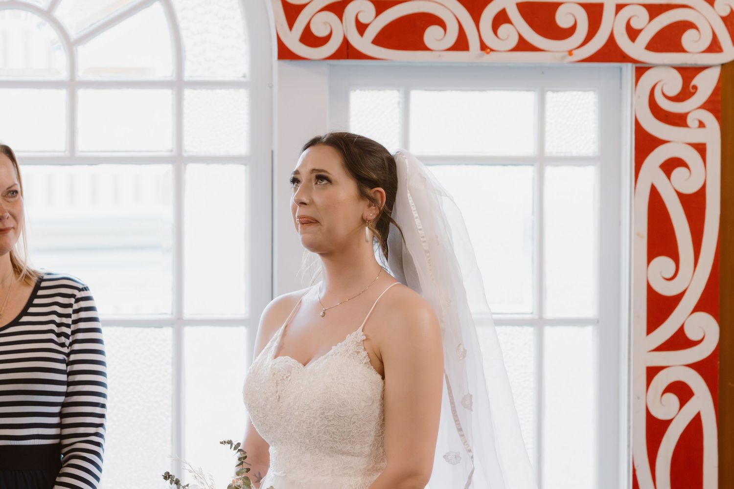Bride in white dress stands by sunlit arched window with decorative red trim.