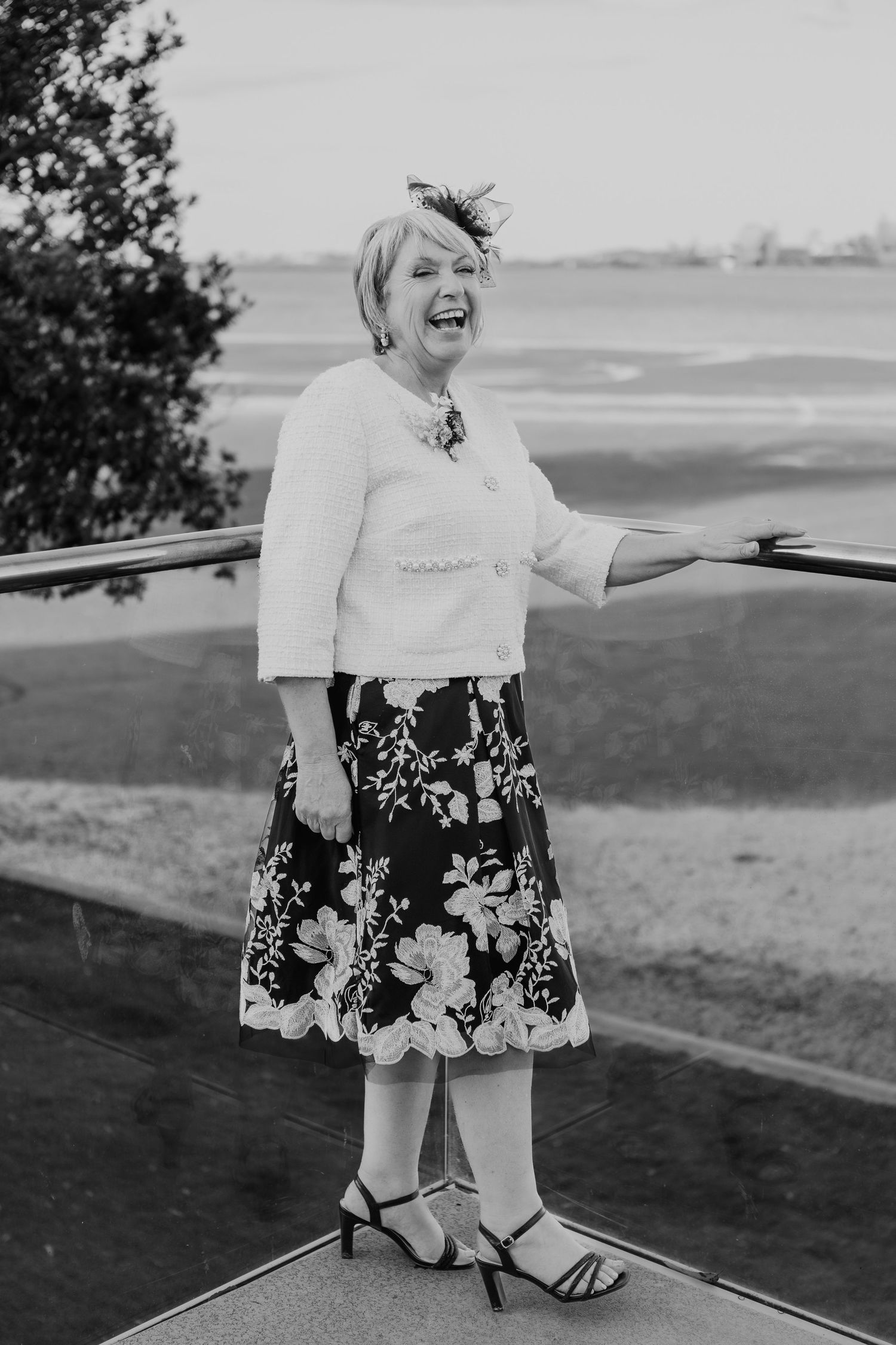 Woman in floral dress and white jacket smiling outdoors at coastal venue.