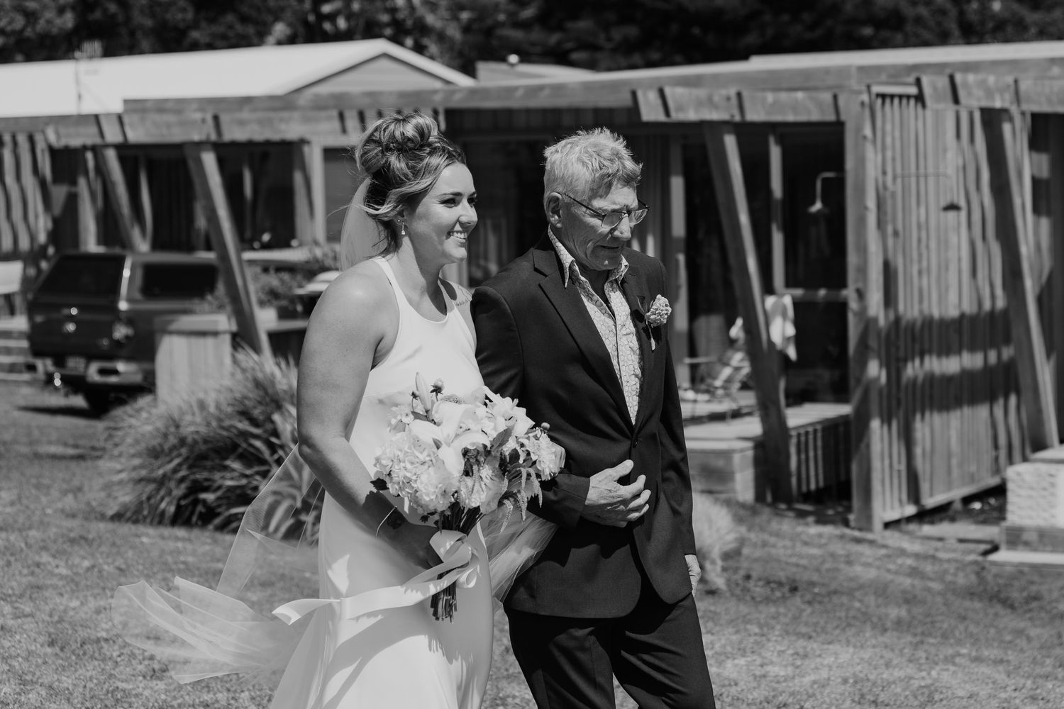 Black and white photo of a bride walking down the aisle with an older man at an outdoor wedding ceremony.