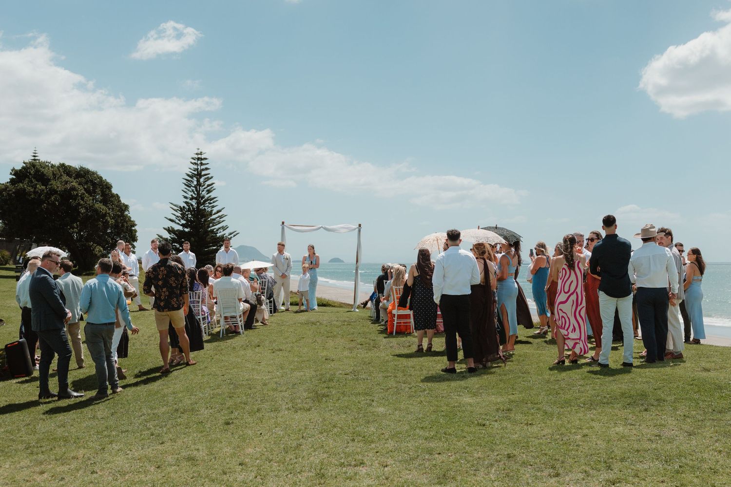 Outdoor wedding ceremony on green lawn by the ocean with guests gathered under blue sky.