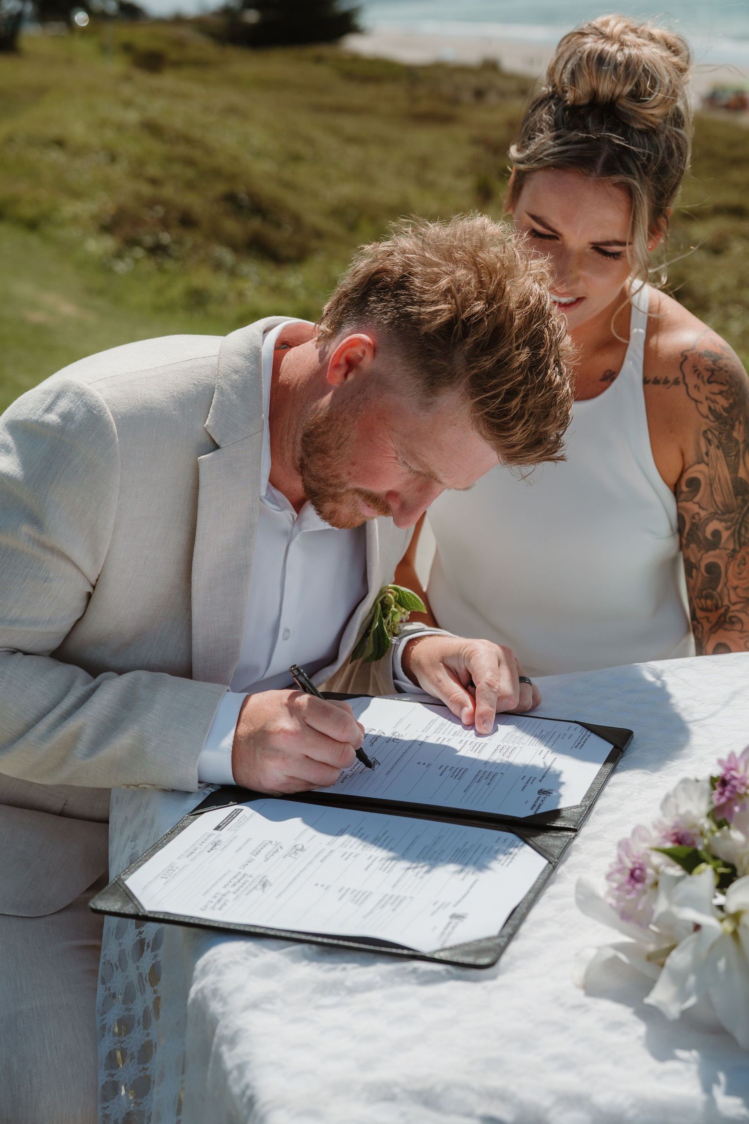 Groom in white suit signing wedding certificate at outdoor table while tattooed bride smiles beside him.