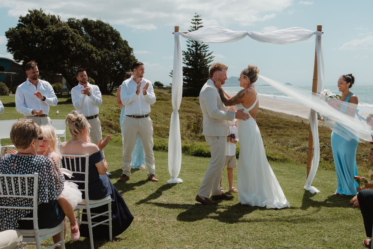 Outdoor wedding ceremony on green lawn with bride and groom embracing under white draped arch, guests seated in white chairs.