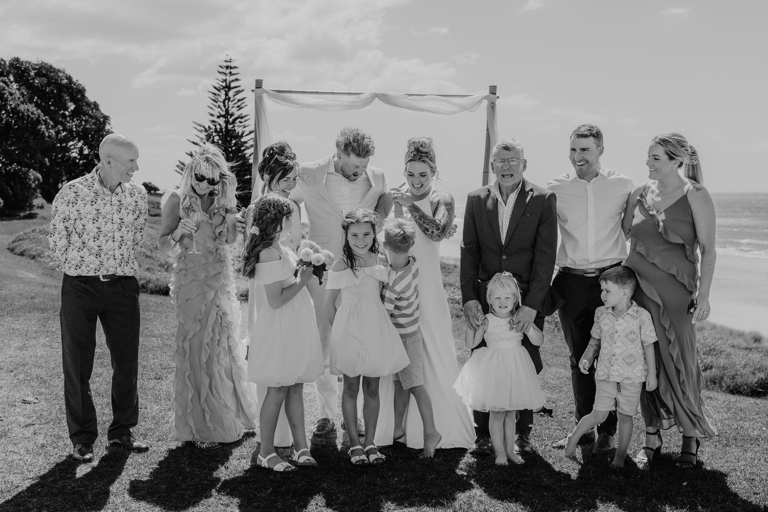 Black and white wedding photo of bride, groom, and guests posing outdoors near the ocean under a floral arch.
