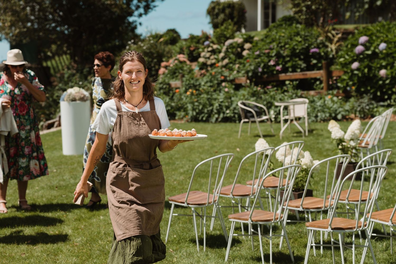 Smiling caterer in brown apron carries appetizer platter at outdoor garden wedding reception with white chairs.