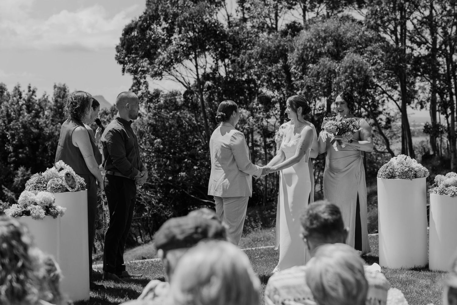 Outdoor wedding ceremony with couple holding hands before officiant, guests seated, lush trees backdrop, black and white photo.