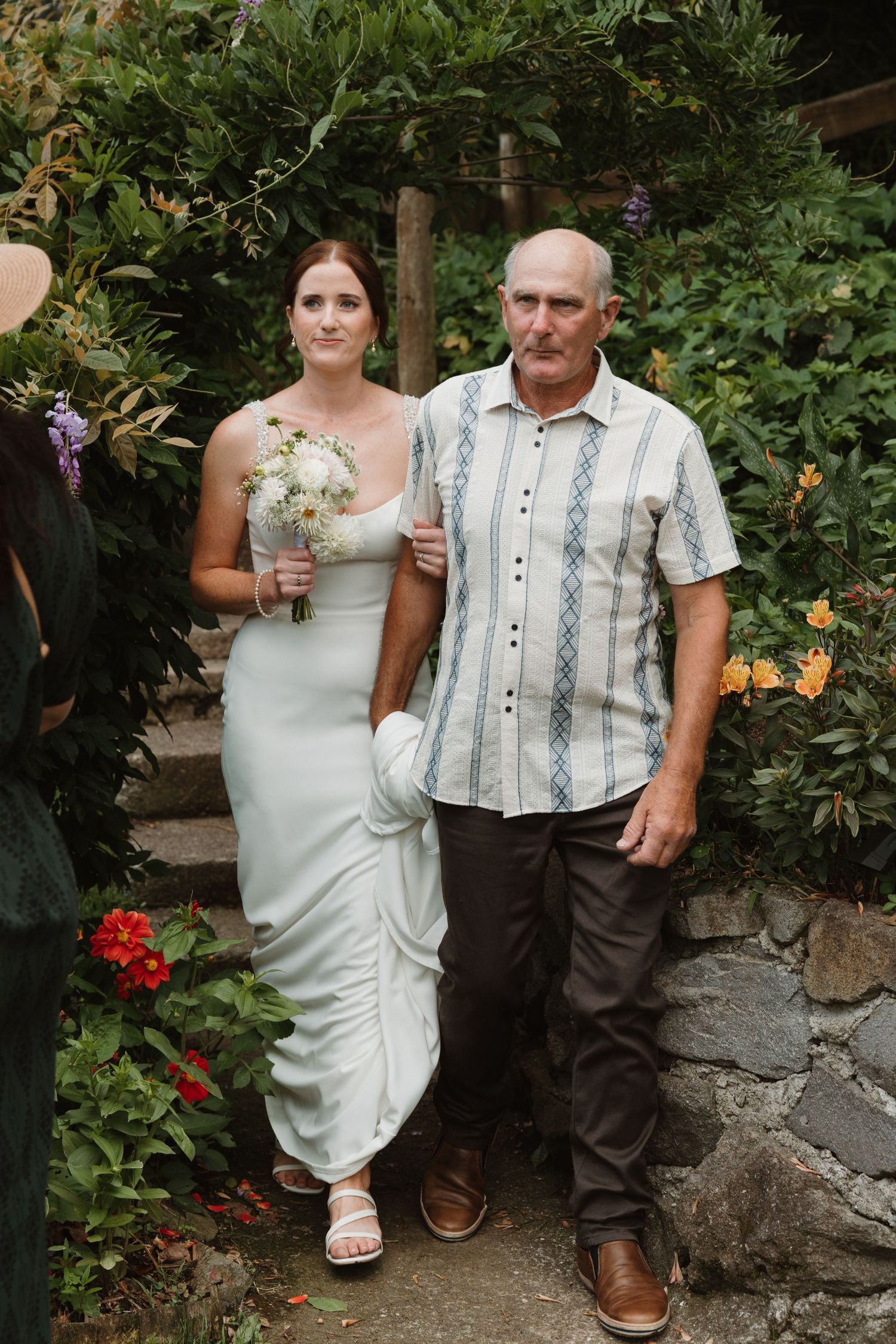 Bride in white gown holding bouquet walks with older man in striped shirt through garden wedding ceremony.