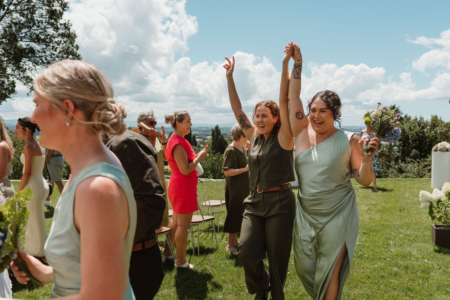 Bridesmaids in sage green dresses celebrate joyfully outdoors at a sunny wedding ceremony with guests in background.